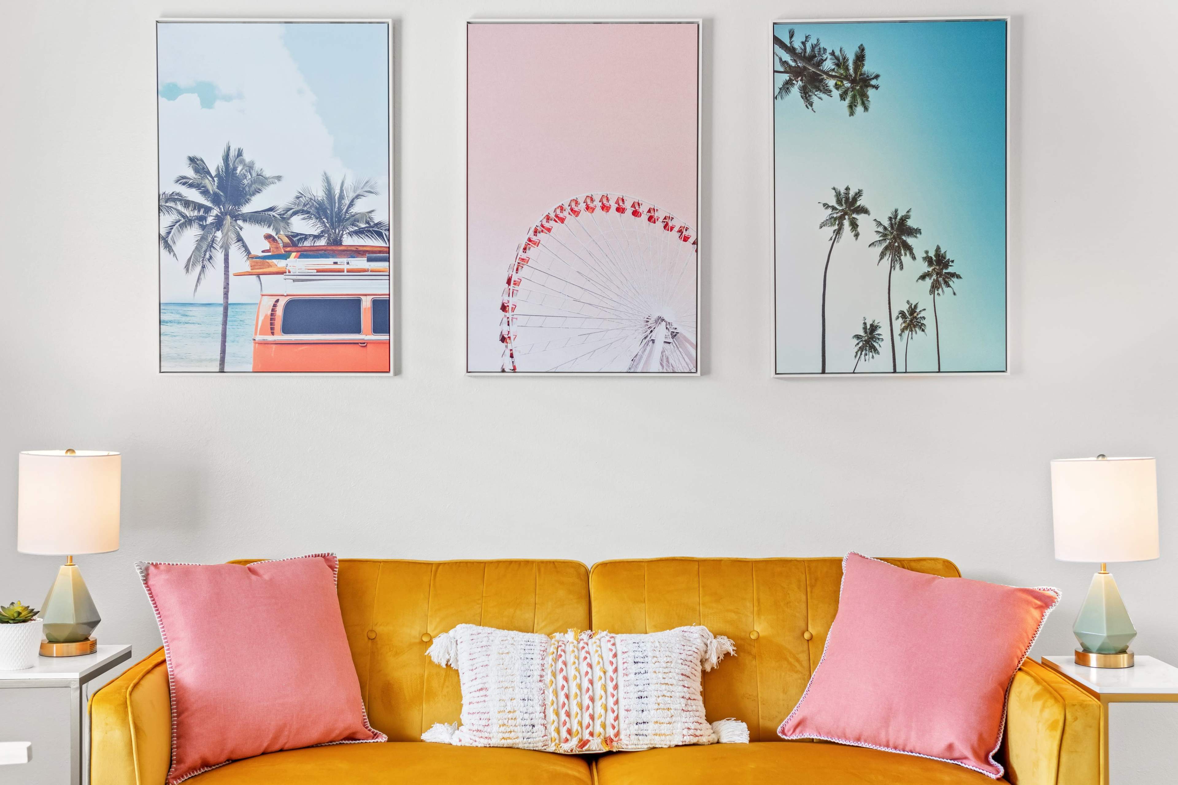 A yellow sofa with pink cushions is positioned below three framed landscape prints featuring a vintage van, a Ferris wheel, and palm trees.