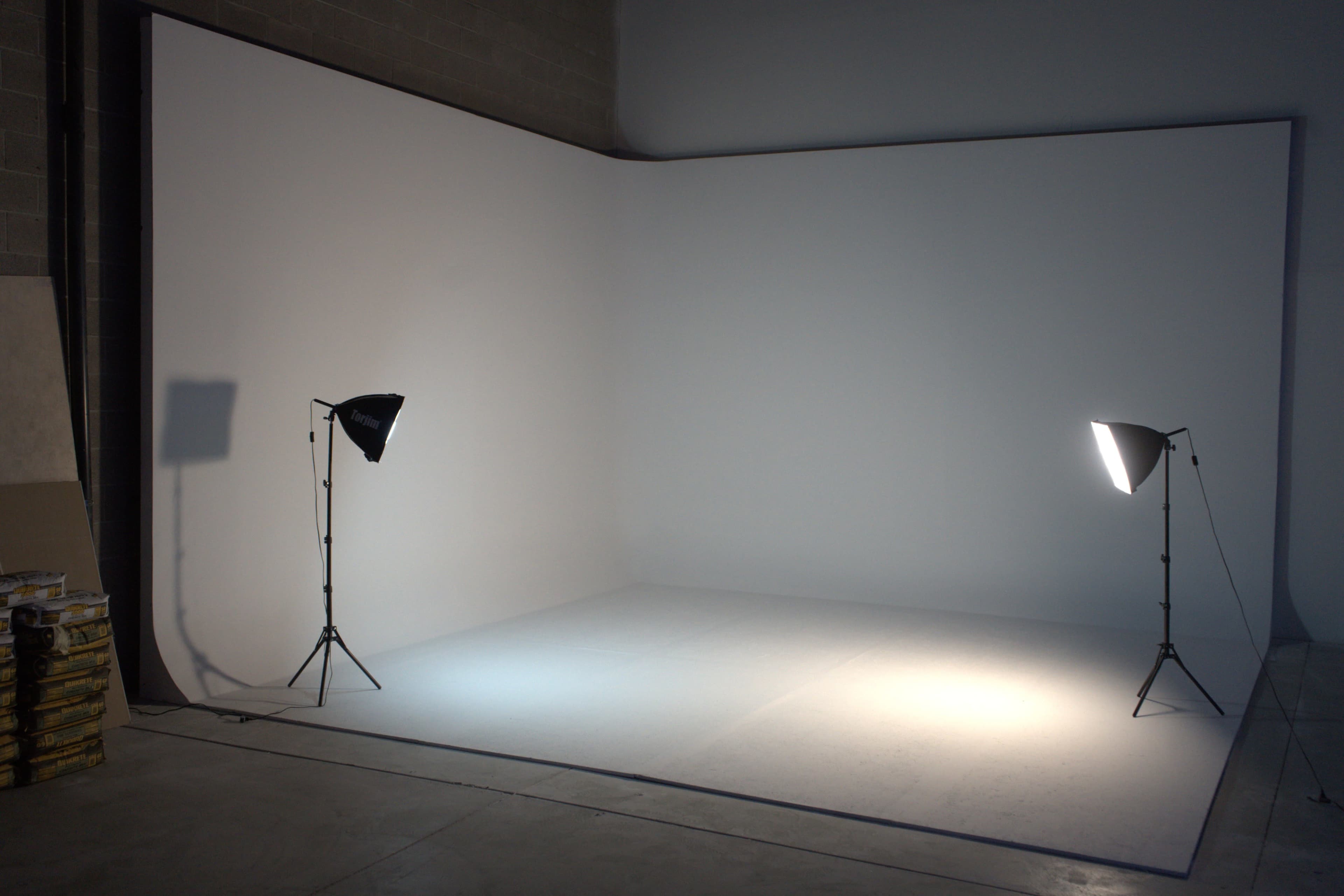 The image shows a minimalist photography studio with a blank white backdrop and two lighting stands positioned on opposite sides.