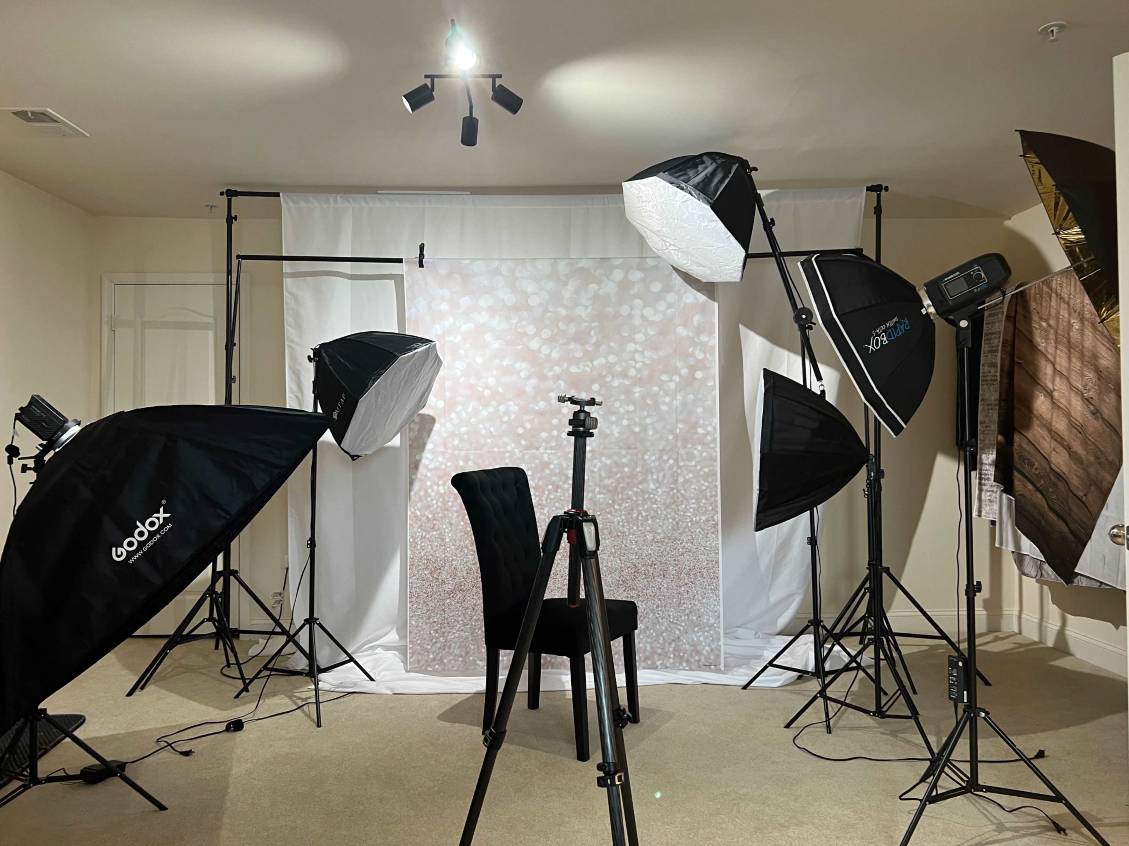 A photography studio setup featuring multiple softbox lights, a backdrop with a glittery design, a tripod, and a single chair positioned in the center.