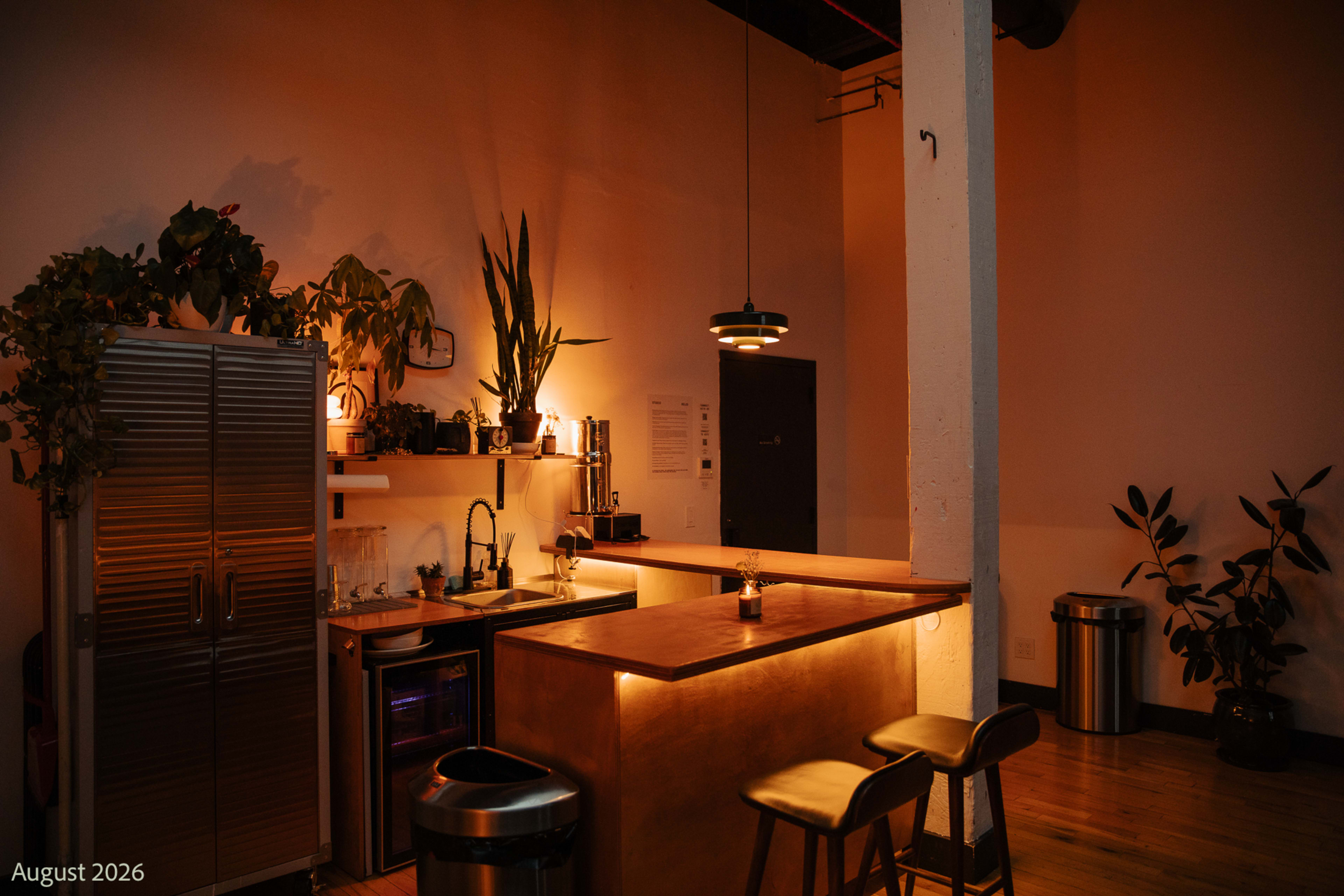 The image shows a modern kitchen area featuring a wooden countertop, bar stools, plants, and stainless steel appliances, with warm lighting creating a cozy atmosphere.