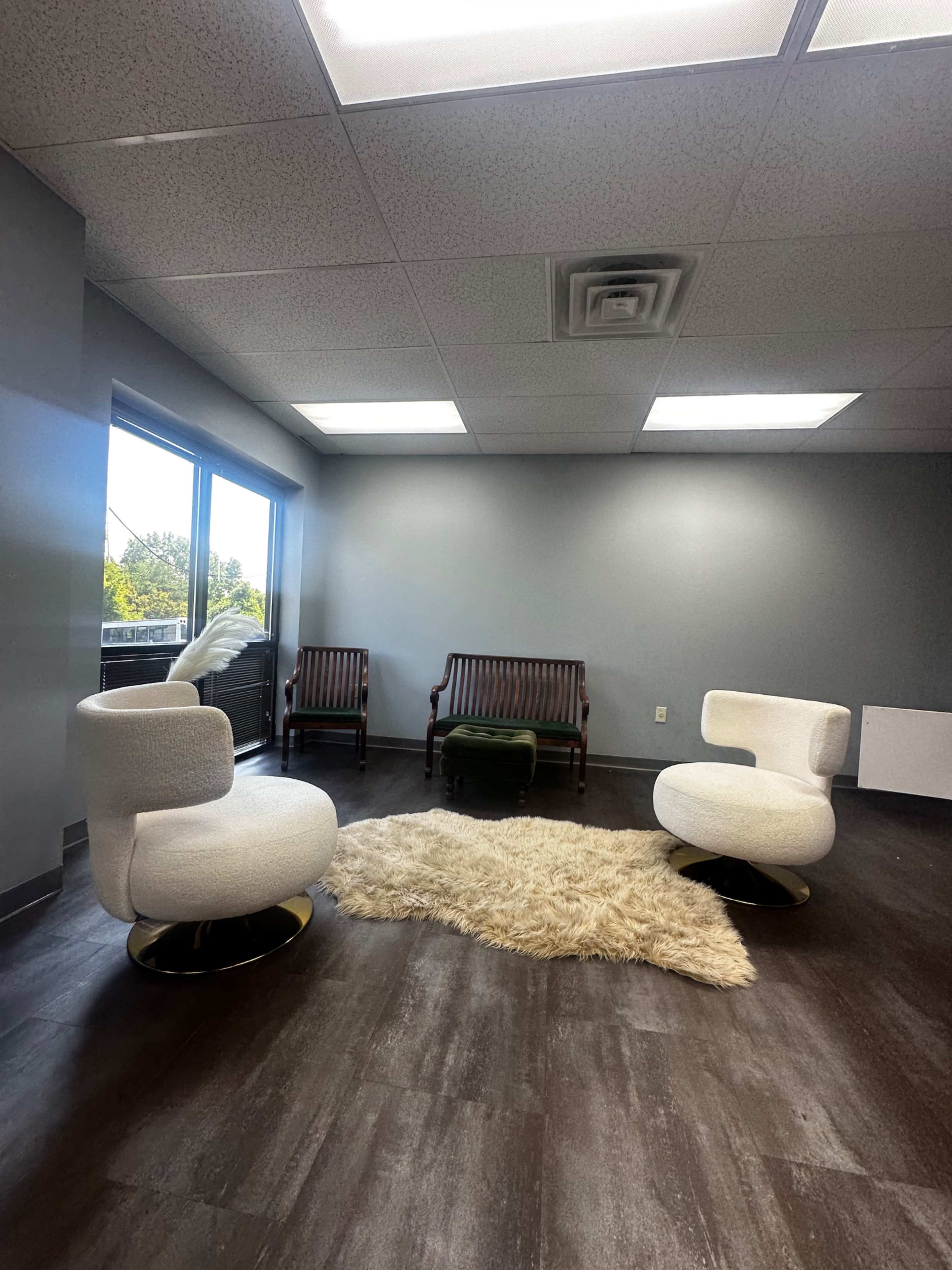 The image shows a modern waiting area with two white swivel chairs, a beige rug, and two wooden benches against a gray wall, illuminated by natural light from large windows.