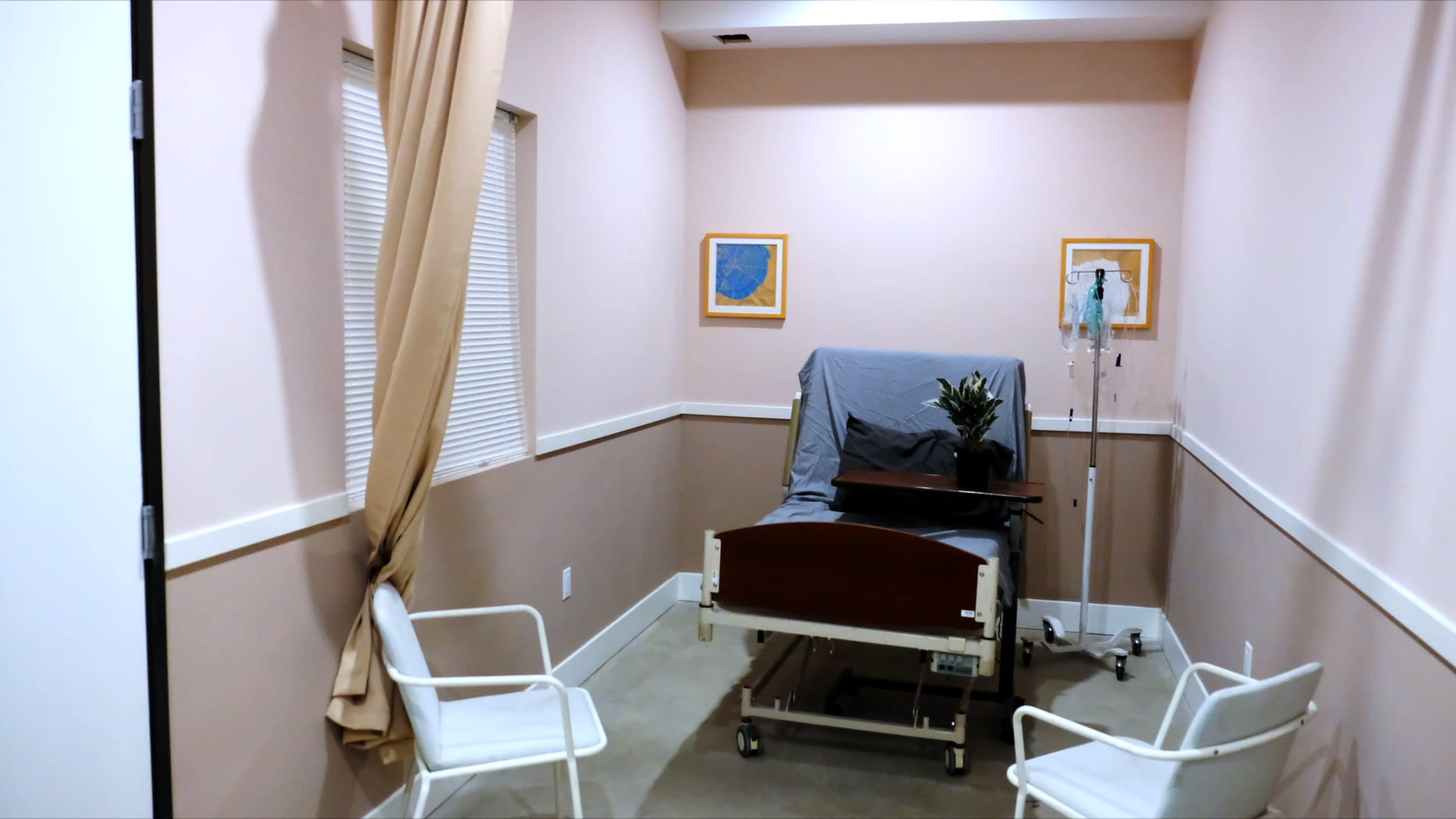 The image depicts a small hospital room featuring a bed with a gray cover, an IV stand, and two white chairs next to a window with curtains drawn.