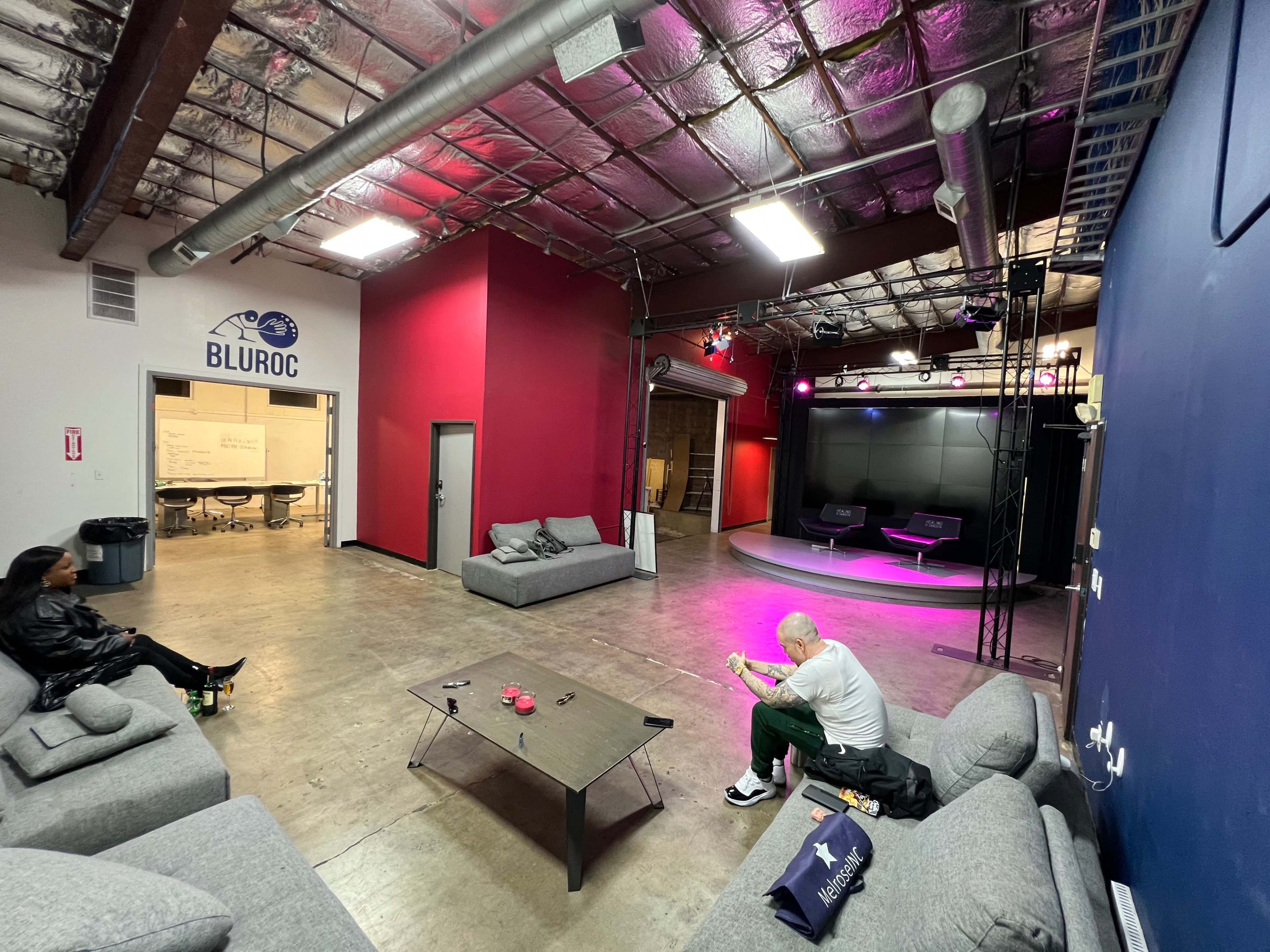 A spacious, industrial-style room features multiple gray couches, a black table, and a small stage area with lighting in the background.