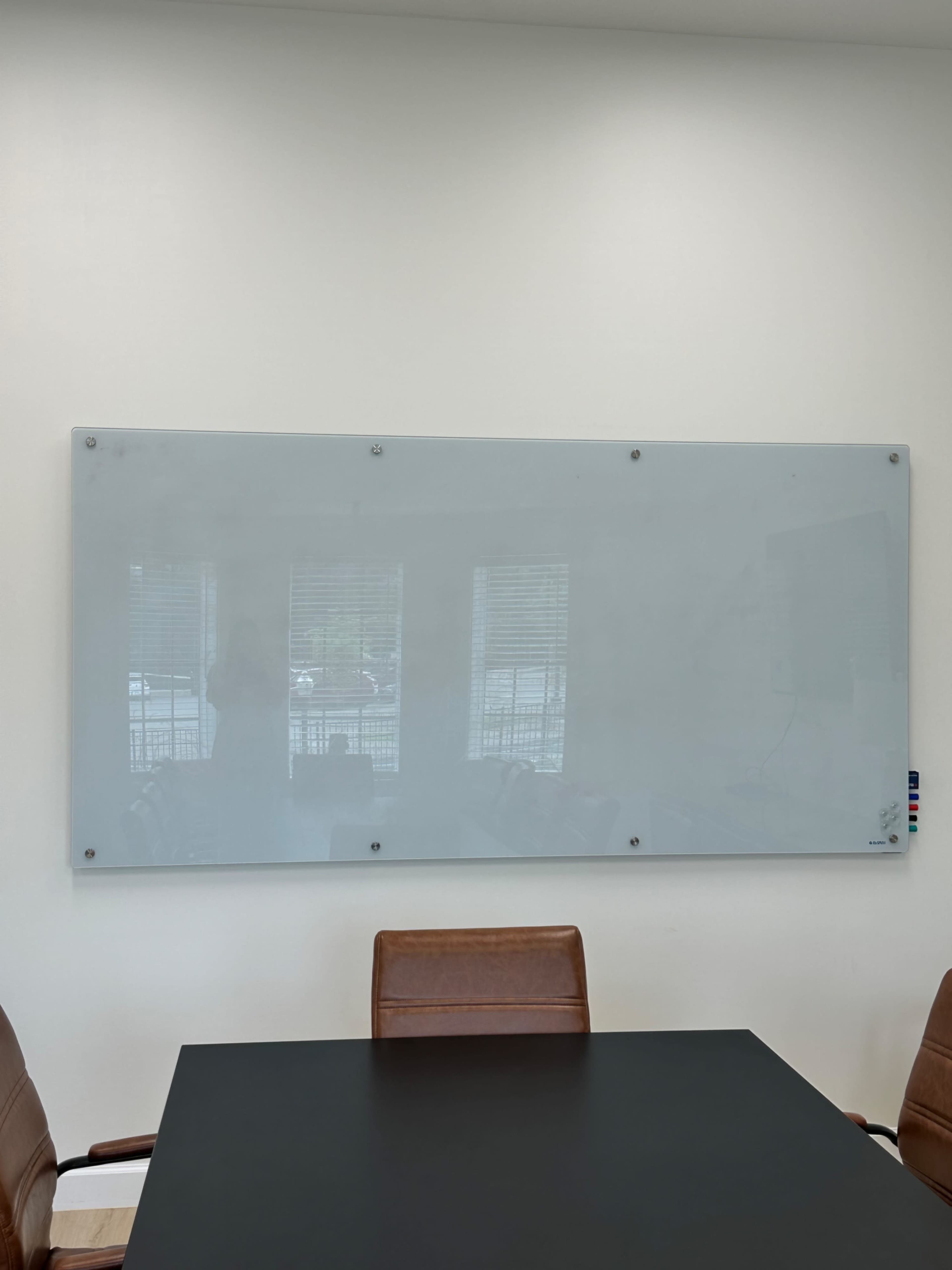 The image shows a glass whiteboard mounted on a wall in a conference room, with a black table and brown chairs visible in the foreground.