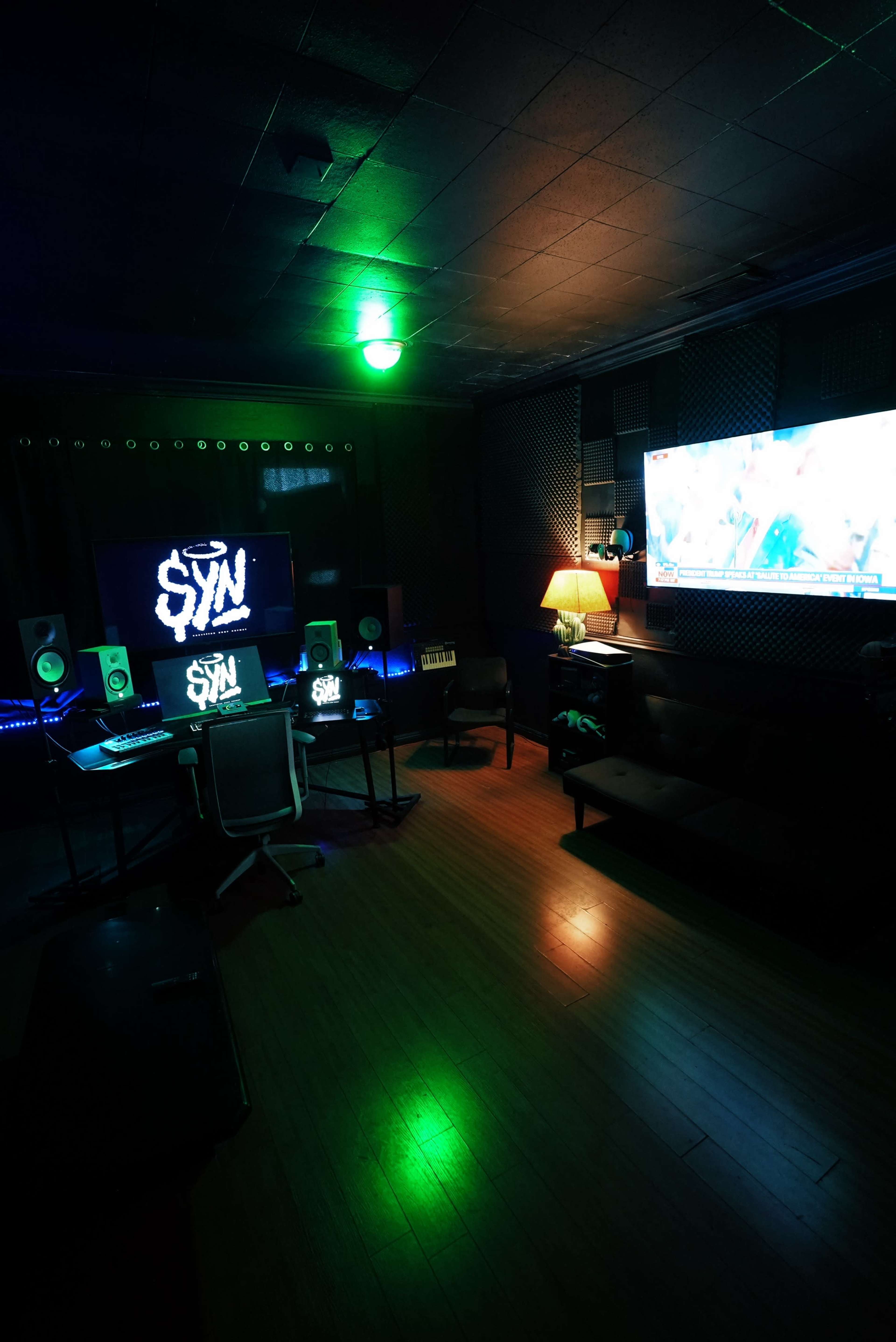 A music production studio features a large mixing desk, speakers, a glowing logo on the wall, and a screen displaying video content, all illuminated by colorful lighting.