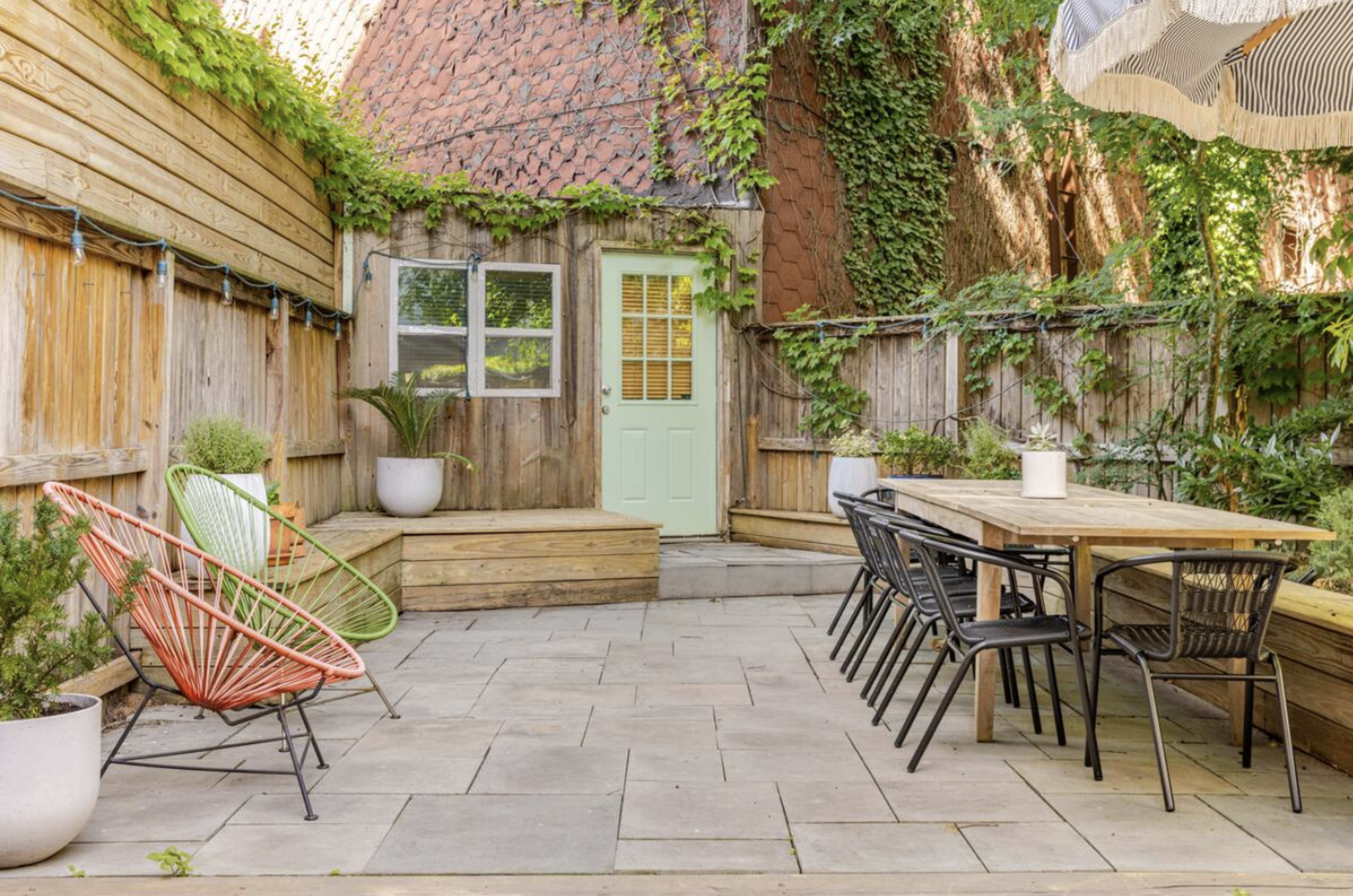 The image shows a landscaped outdoor patio with a wooden table, black chairs, and colorful accent chairs surrounded by greenery and a rustic wooden fence.