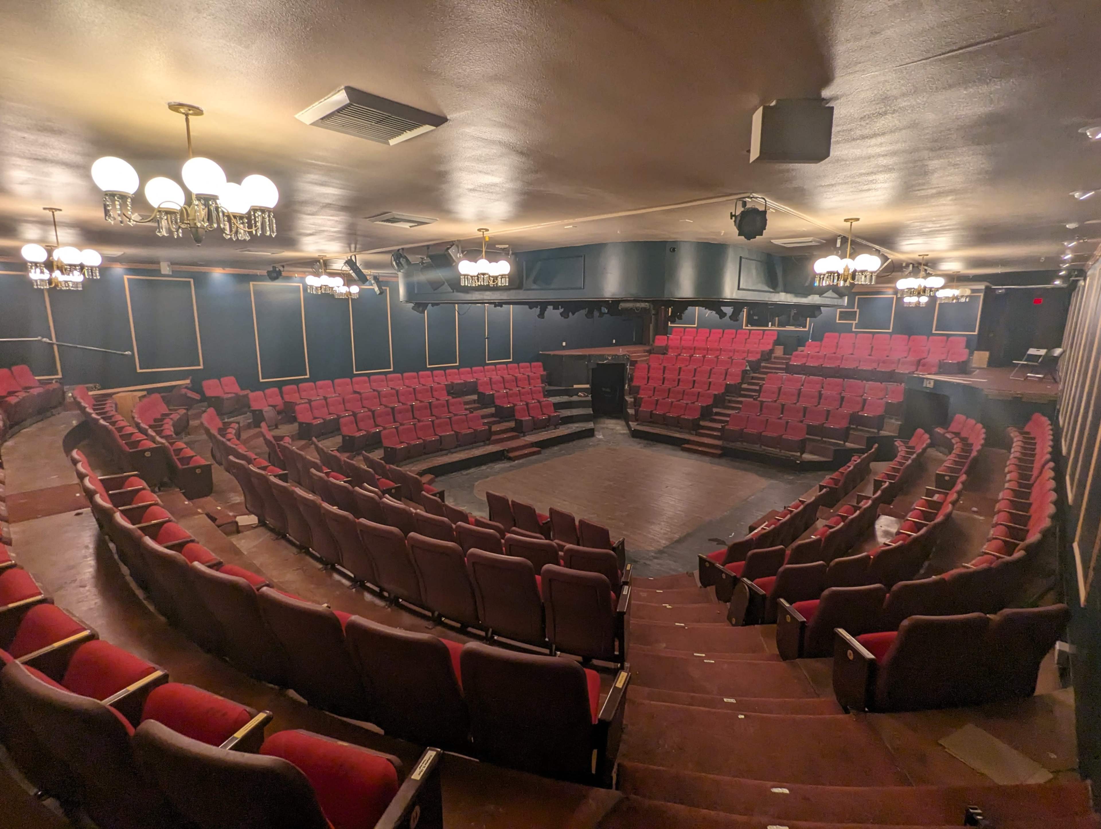 The image shows a circular theater with red seating arranged in a tiered formation around a central stage area, illuminated by overhead lights.