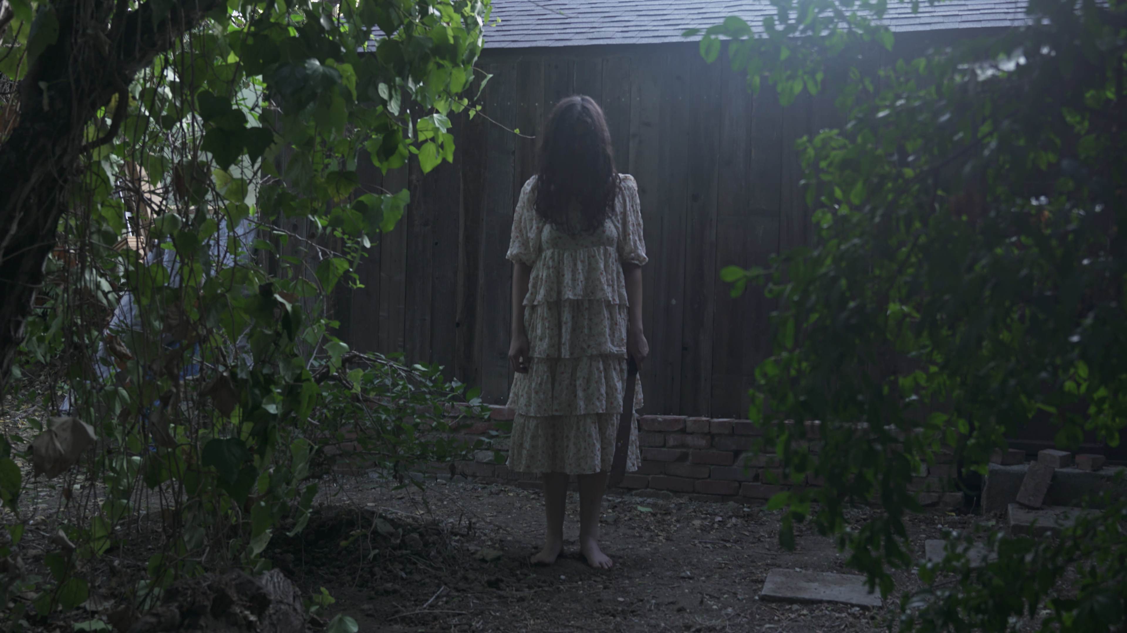 A girl in a white dress stands barefoot in a shaded garden area, surrounded by tall plants and facing a wooden structure.