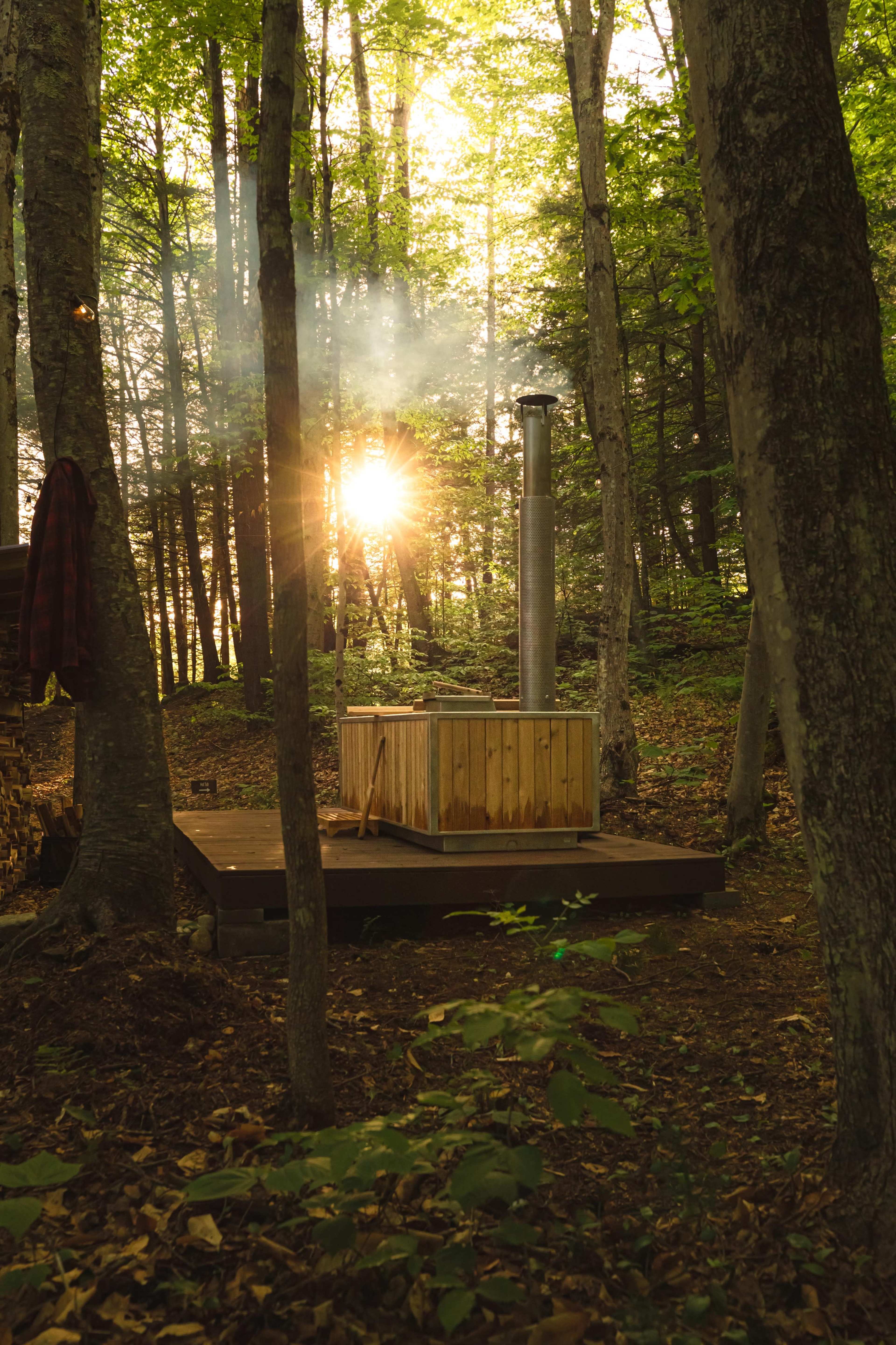 A wooden hot tub emits steam in a forested area as sunlight filters through the trees.