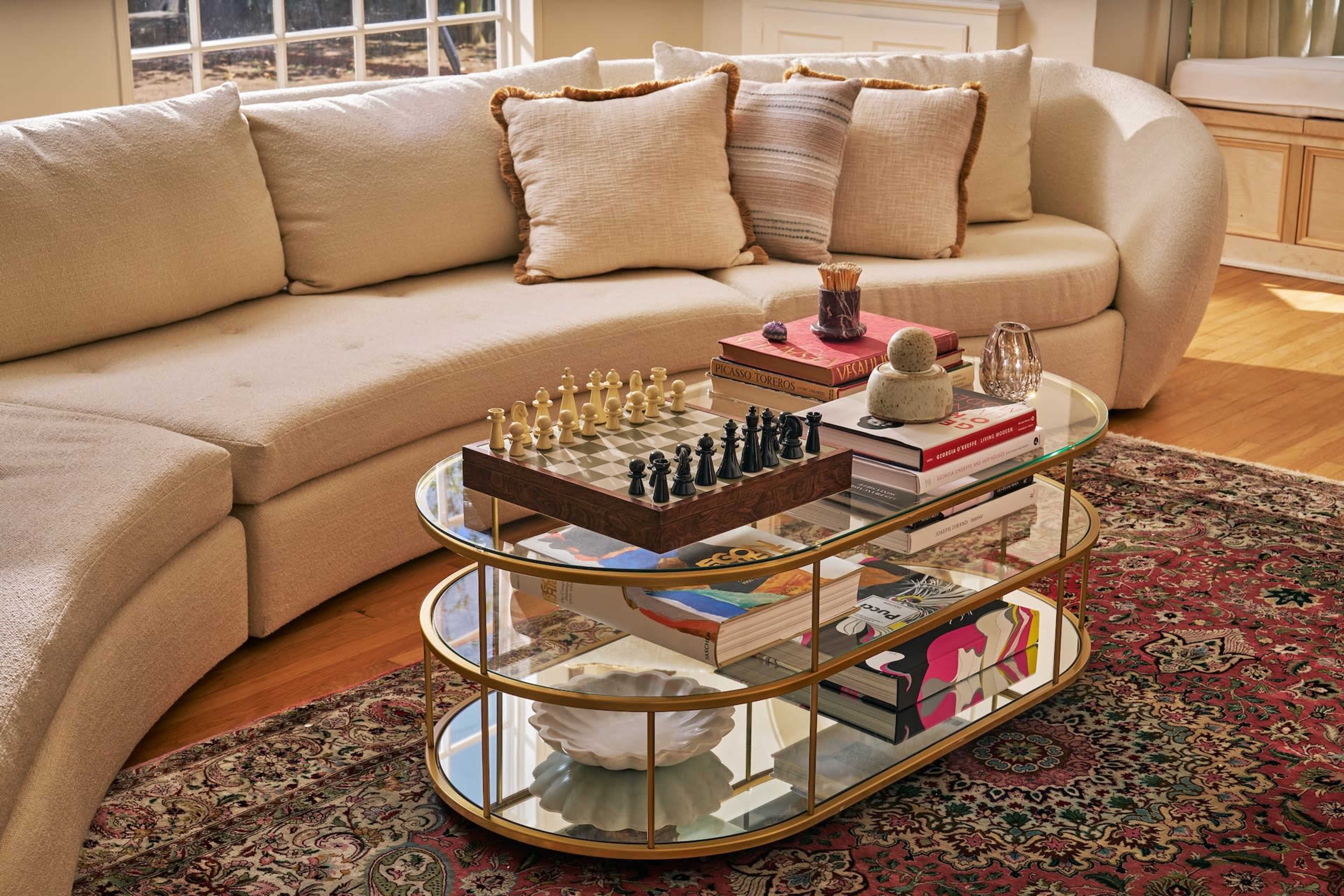 A curved beige sofa with decorative pillows surrounds a glass coffee table, which holds a chess set, books, and a decorative bowl, all on a patterned area rug.