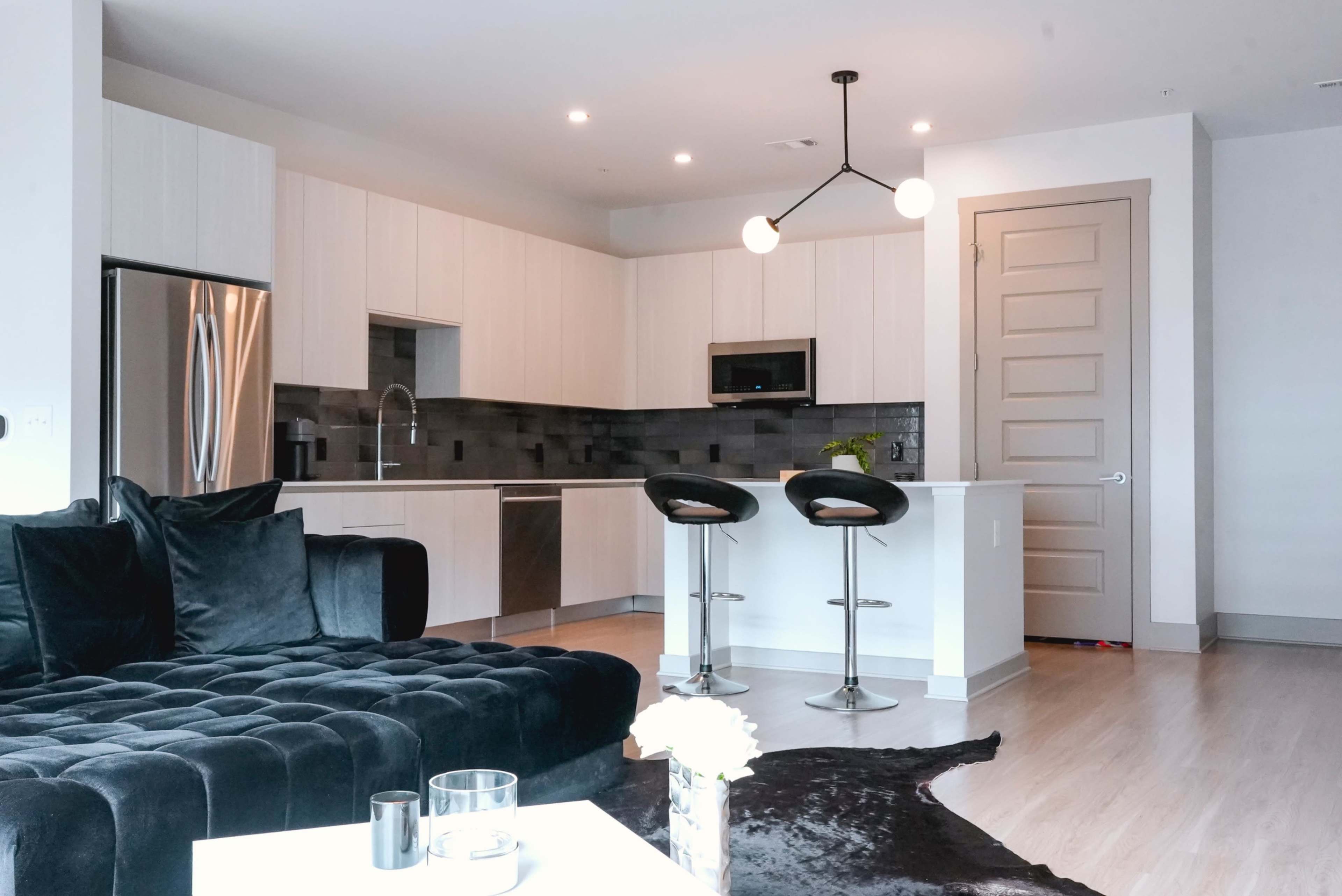 A modern kitchen with light wood cabinets, stainless steel appliances, and a black sectional couch in an open living space.