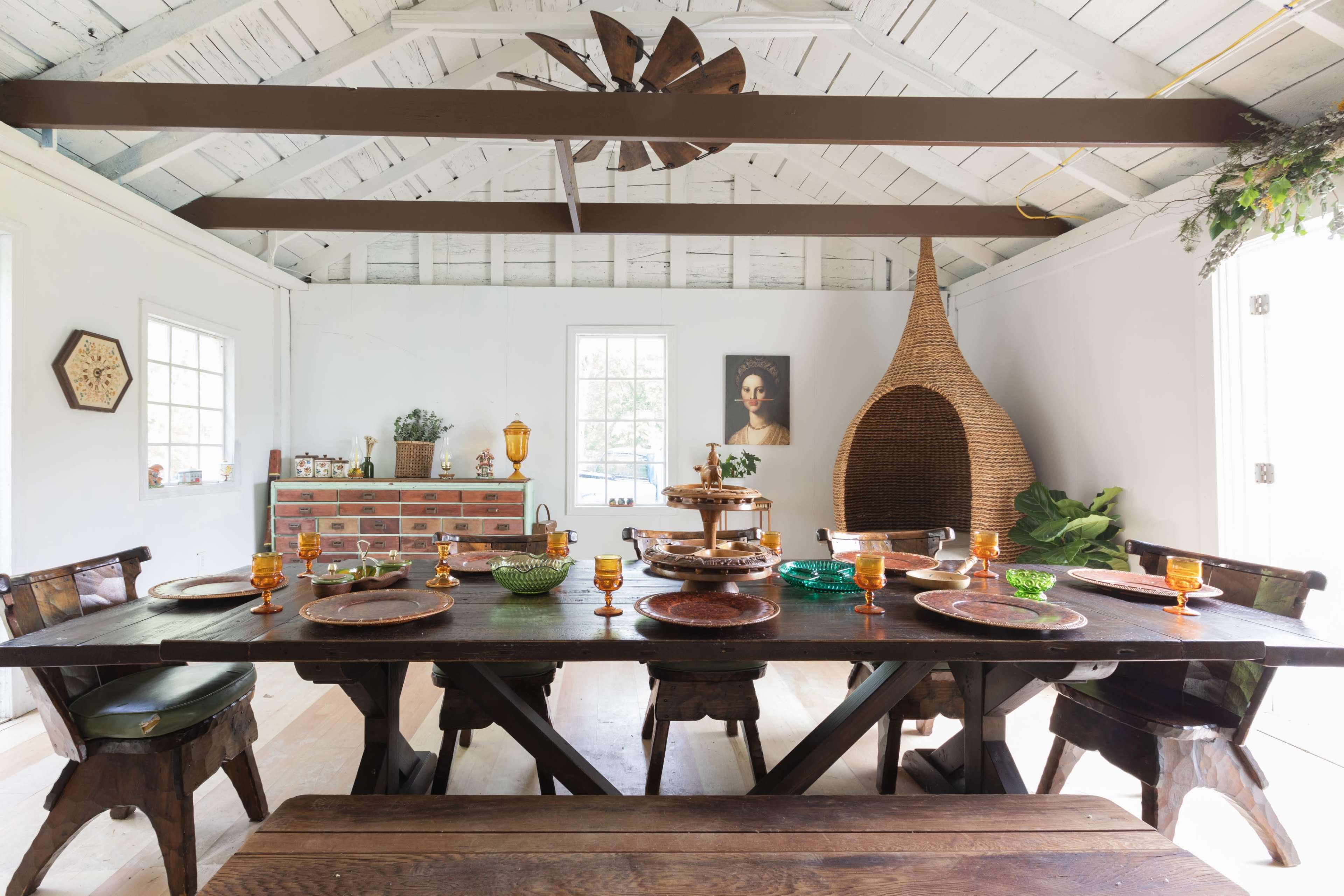 The image shows a spacious dining area with a large wooden table set for a meal, featuring various dishes and colorful glassware, along with a woven chair in the corner and art on the walls.