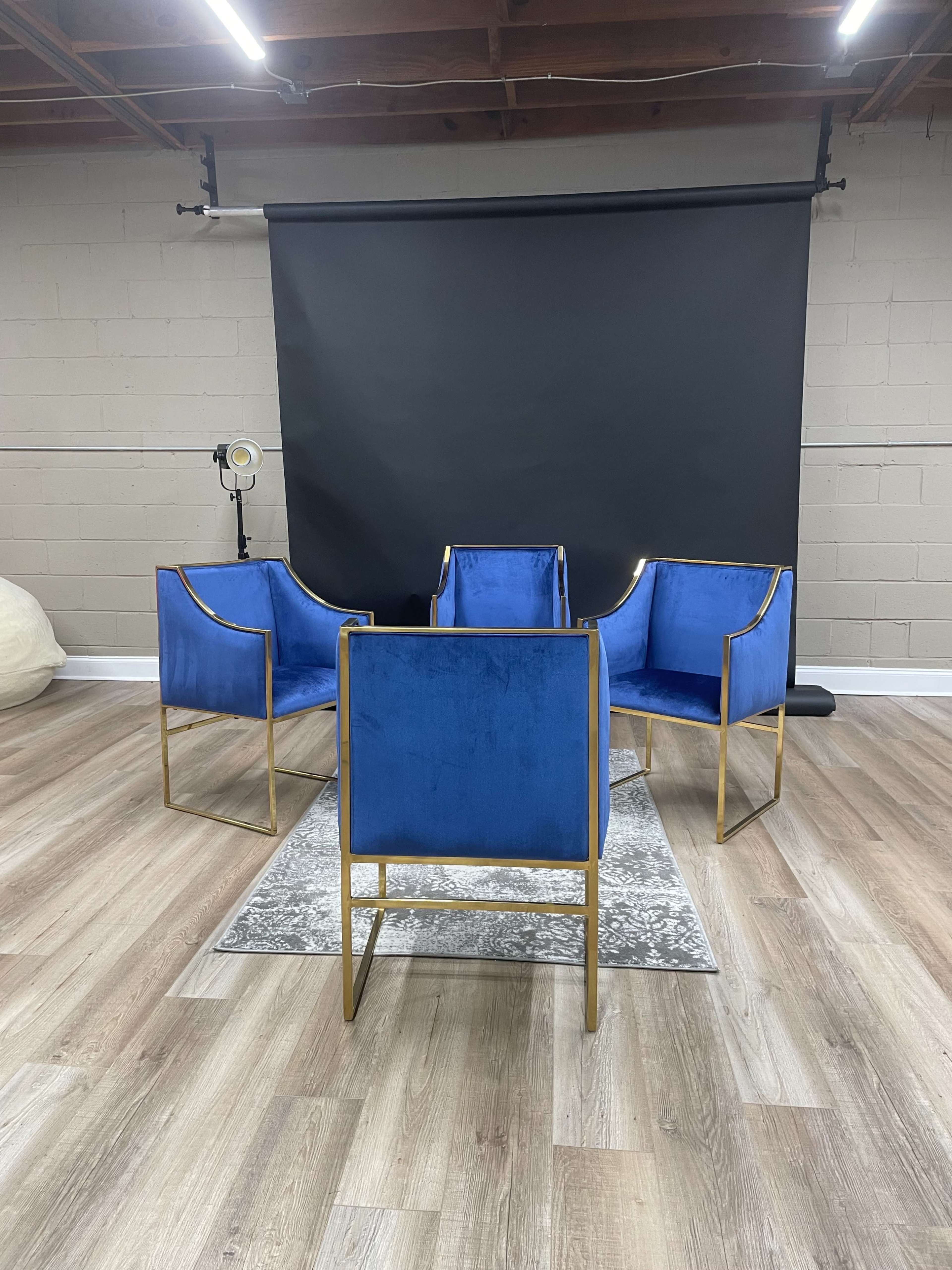Four blue chairs with gold frames arranged around a patterned rug in a room with a black backdrop.