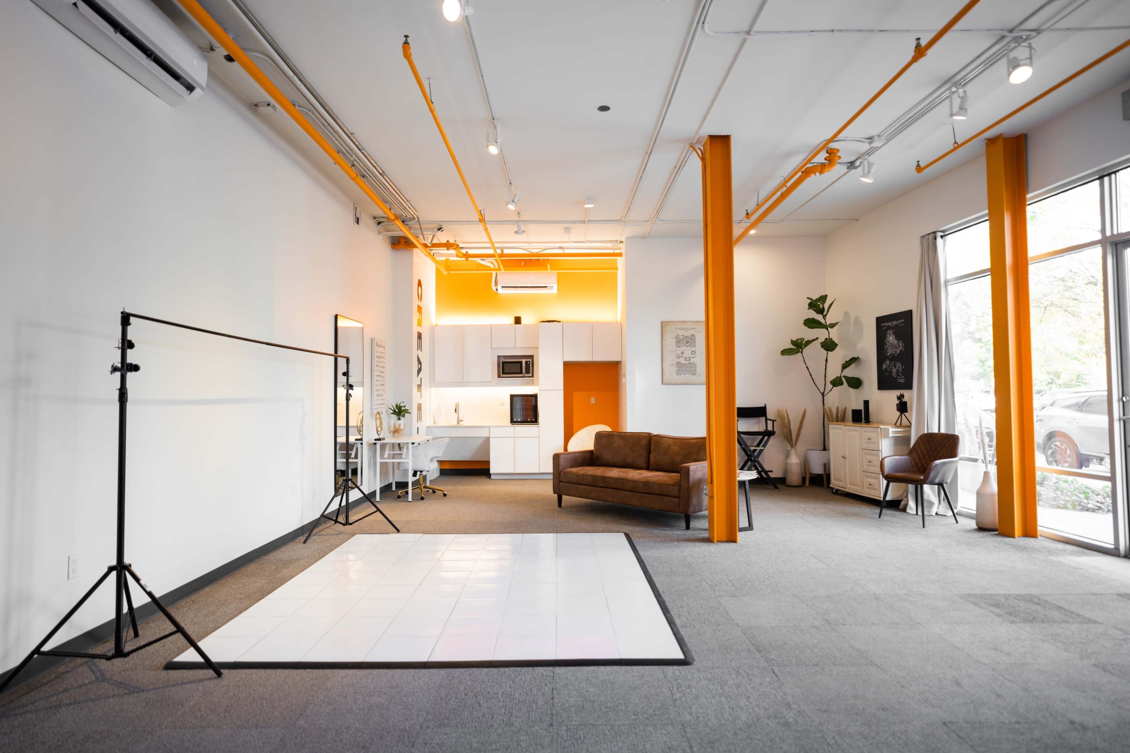 A modern, spacious studio featuring a kitchen area, a brown couch, a white backdrop supported by a stand, and large windows allowing natural light to enter.