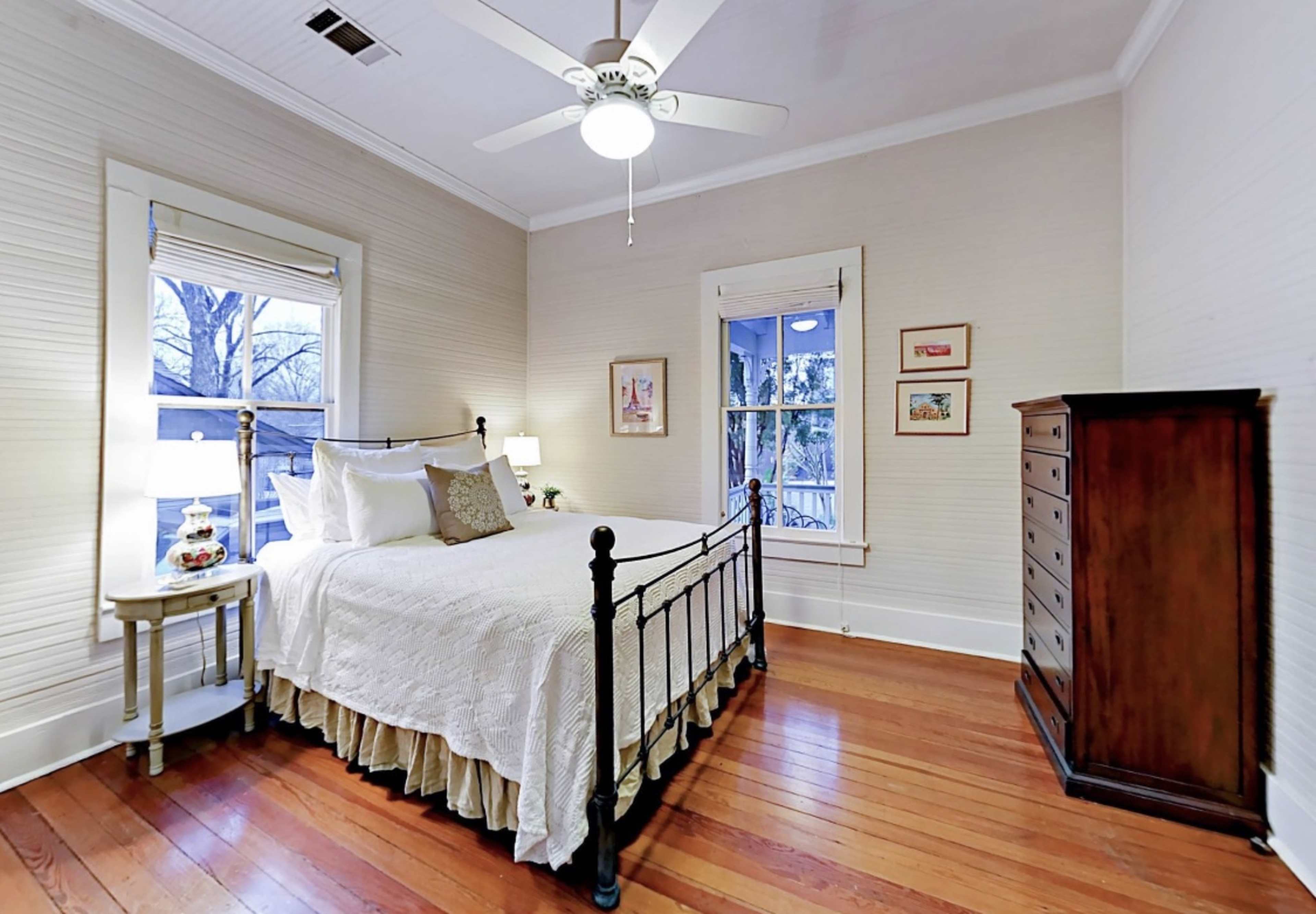 A well-lit bedroom featuring a metal bed frame, a nightstand with a lamp, and a wooden dresser against a wall with beige paneling.