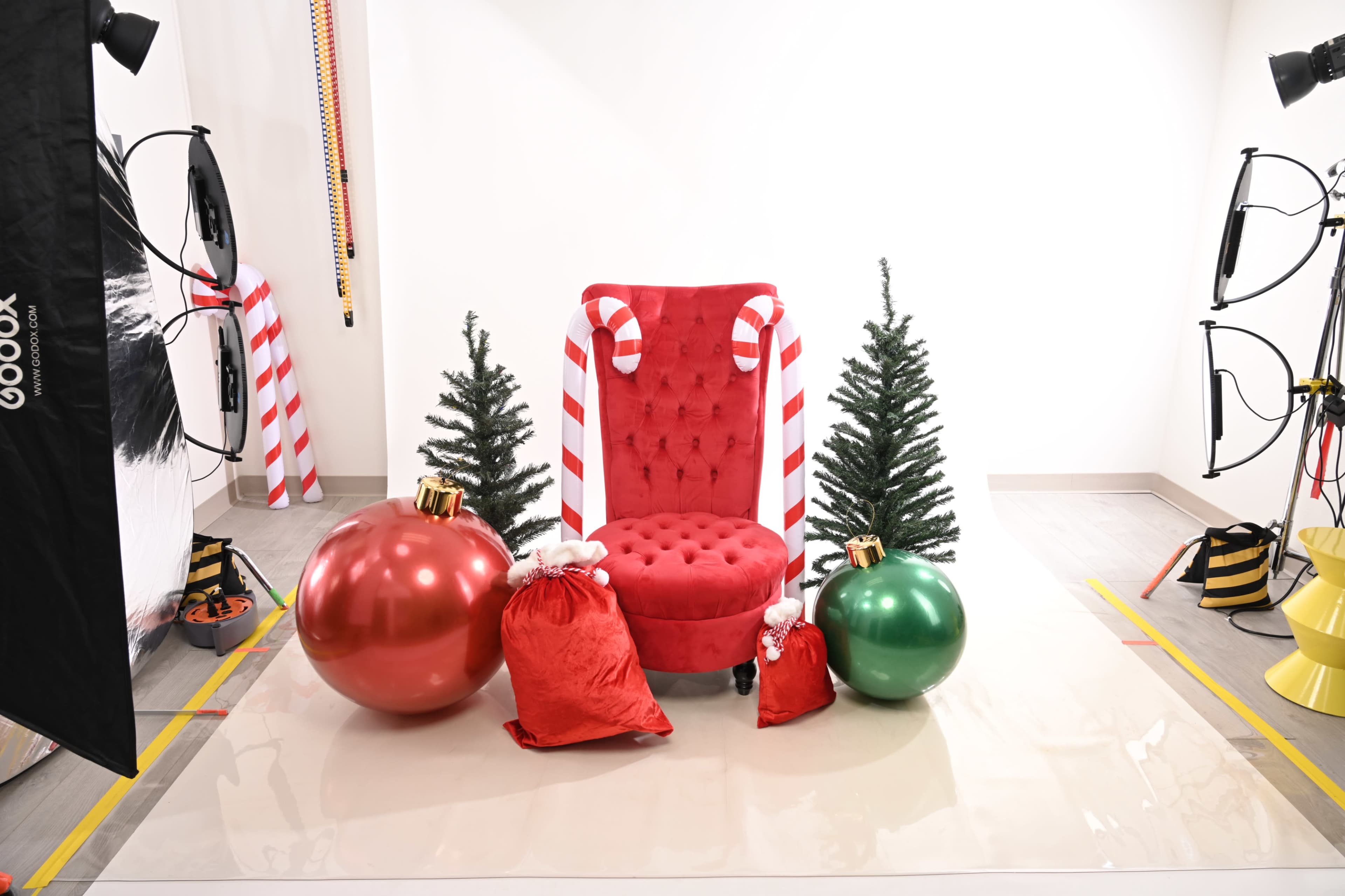 The scene features a red tufted chair surrounded by two small evergreen trees, large red and green ornaments, and red gift sacks, all set in a studio with lighting equipment.