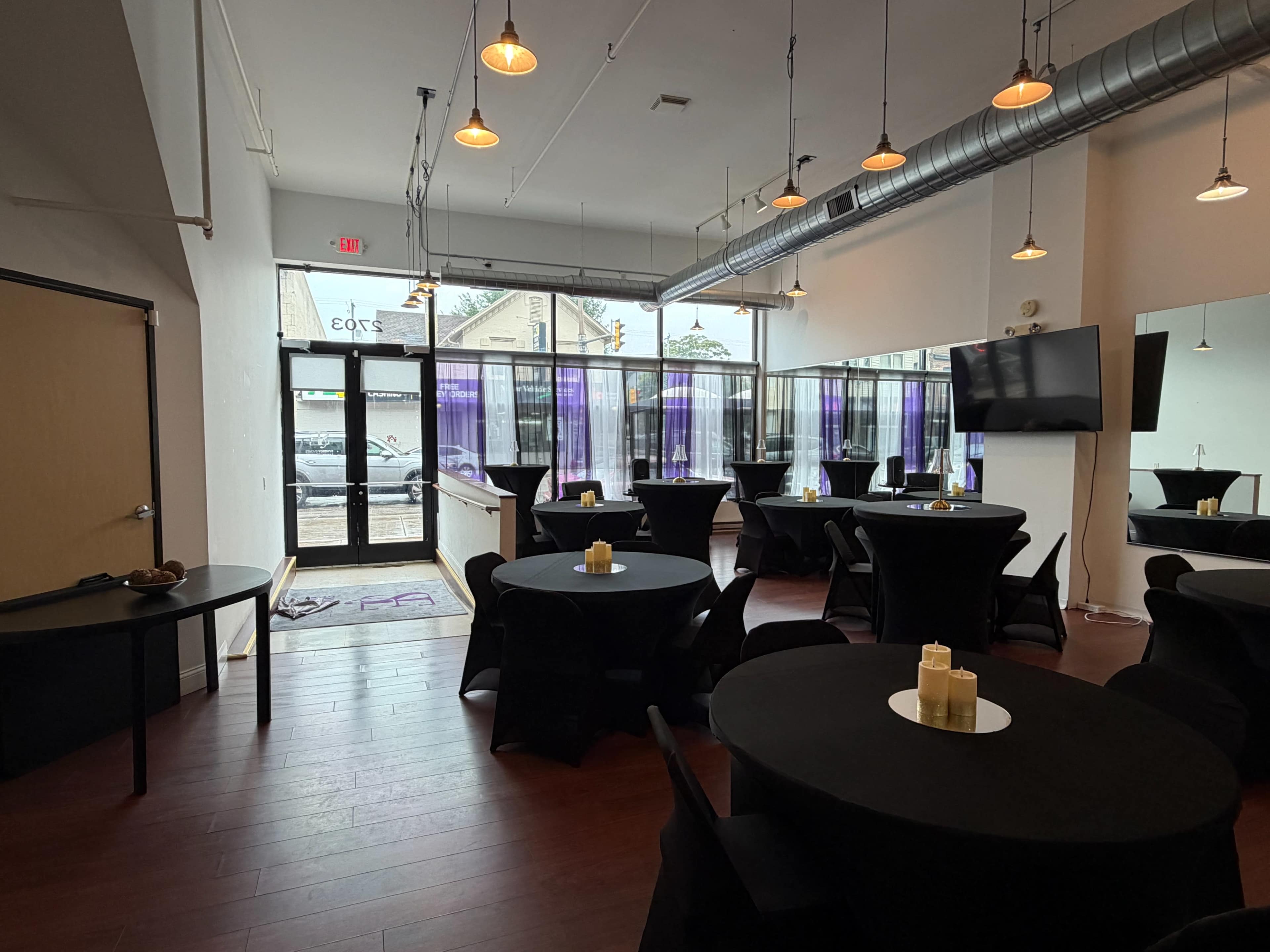 The image shows an interior space with several round tables covered in black tablecloths, featuring candles as centerpieces, and large windows allowing natural light to enter.