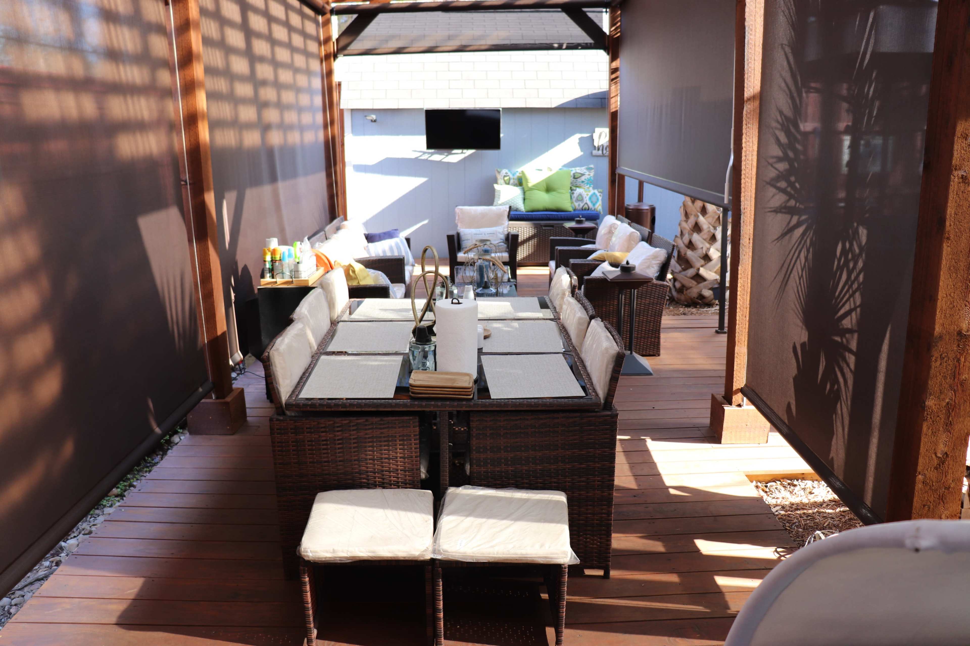 A wooden deck area featuring a sectioned dining table surrounded by wicker chairs, with additional seating and a television mounted on the wall in the background.