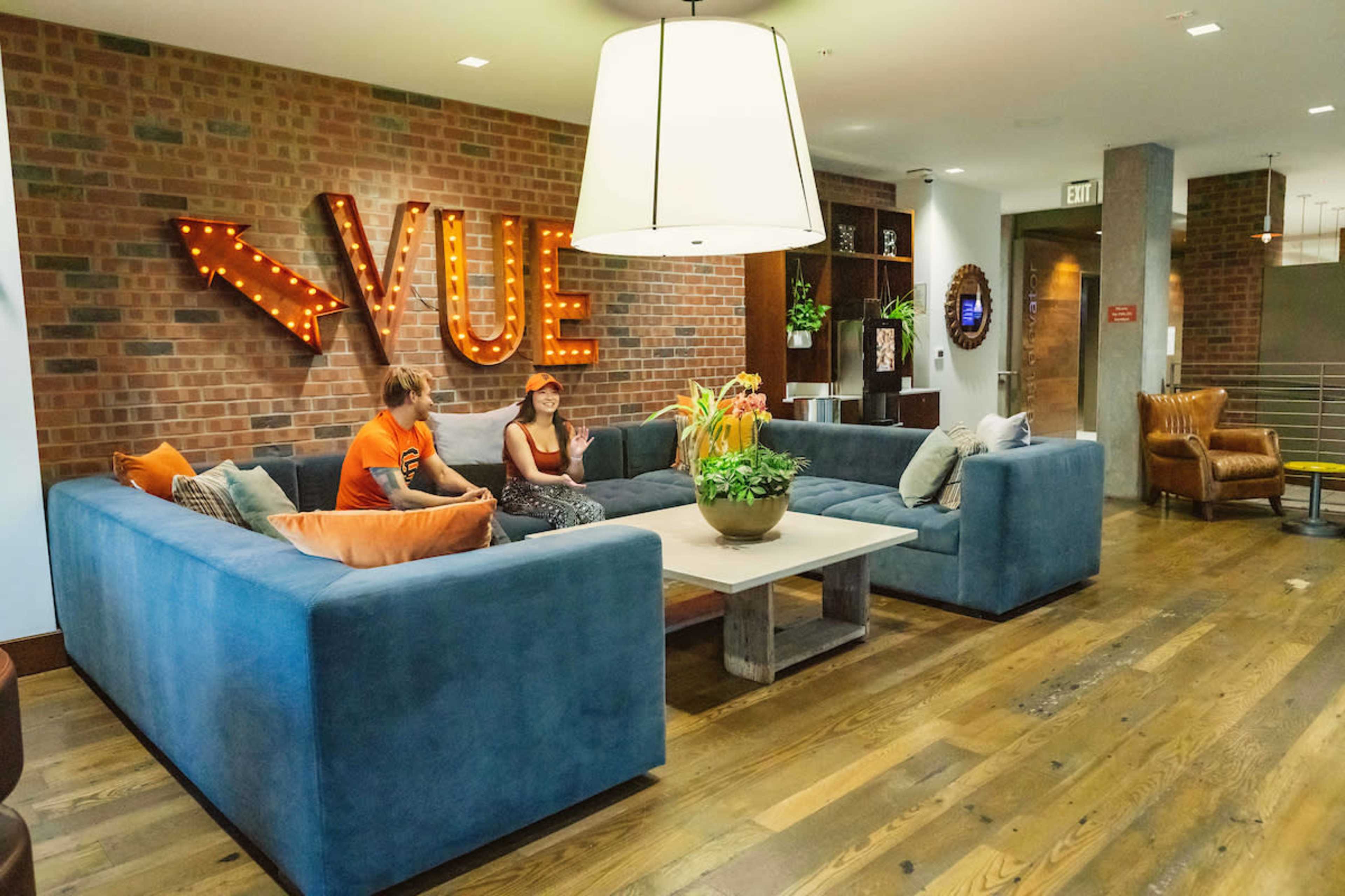 A modern lounge area with a large blue sectional sofa, a coffee table, and decorative lighting, featuring the word "VUE" illuminated on the brick wall.