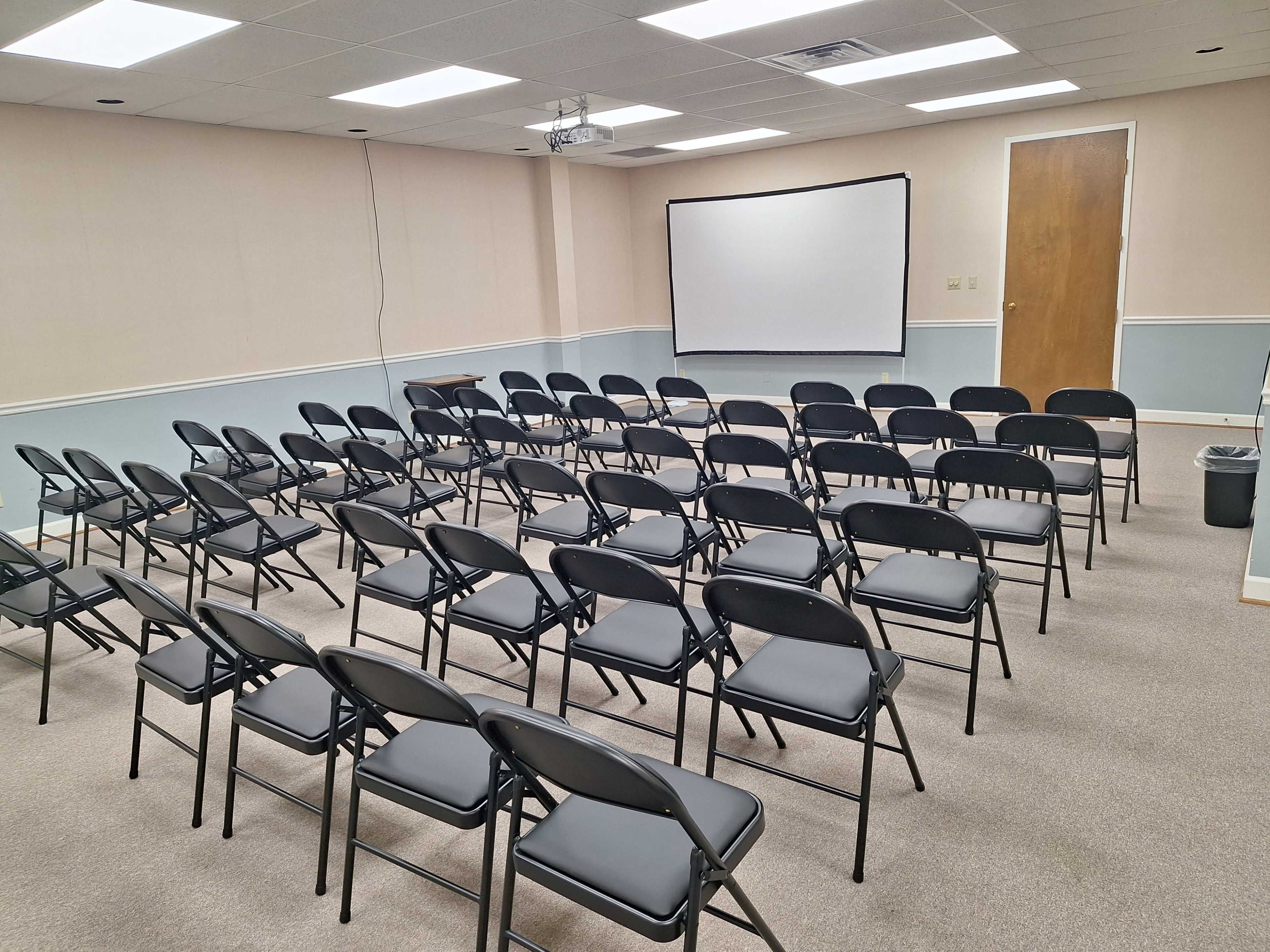 The image shows a meeting room arranged with rows of black folding chairs facing a blank white projection screen.