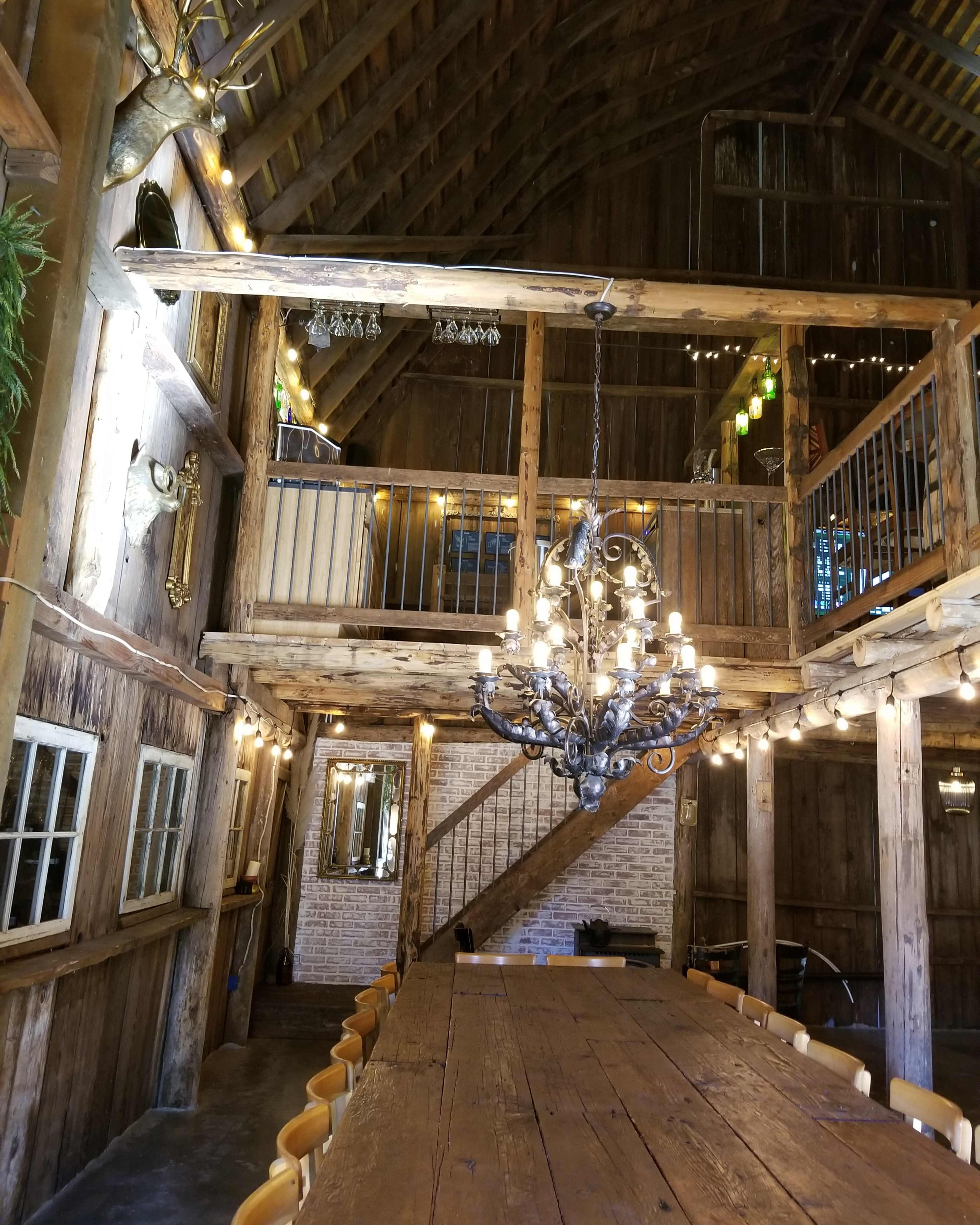 The interior of a rustic barn features a long wooden table, a chandelier, and a staircase leading to an upper level.