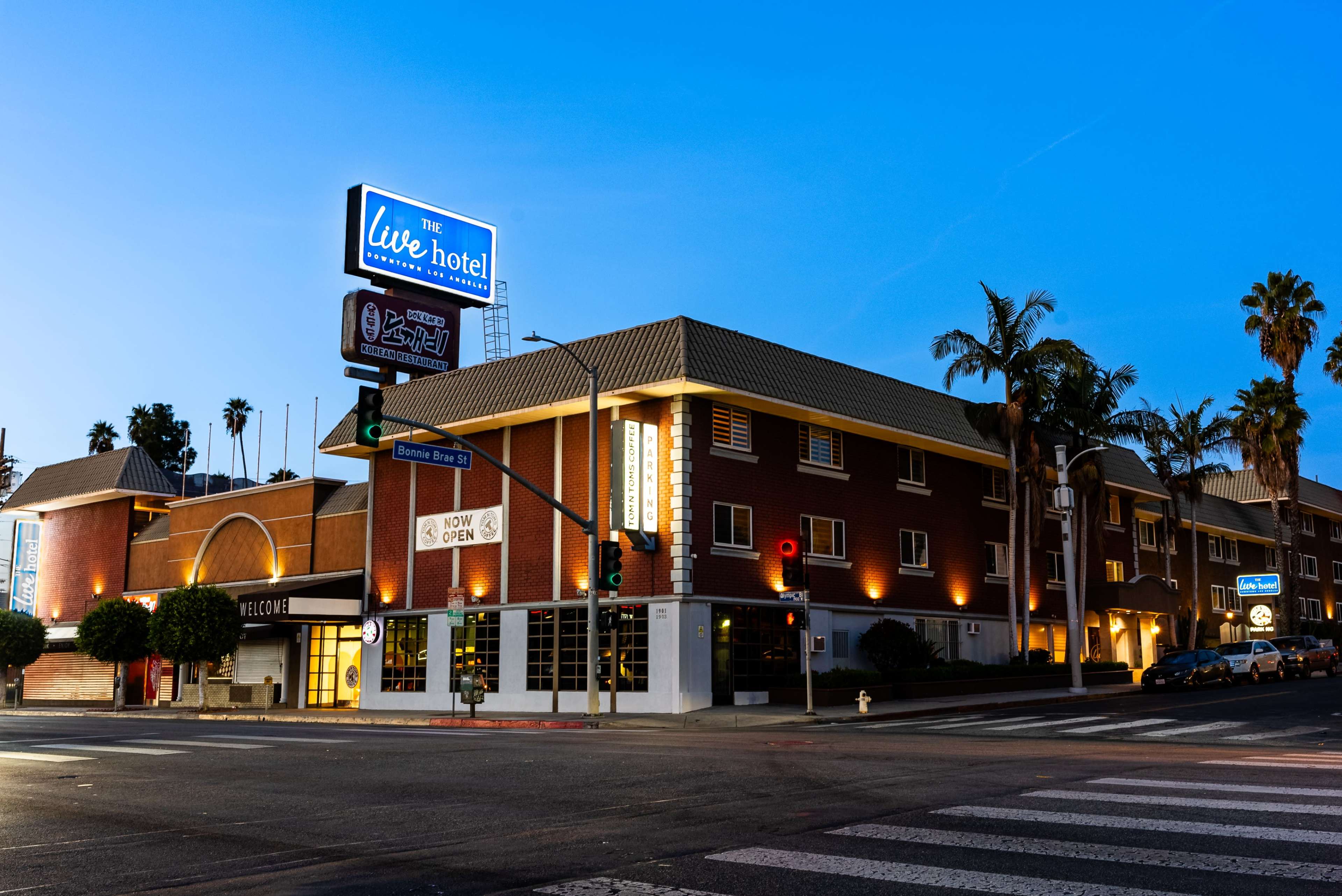 A brick hotel building at dusk, with illuminated signs and palm trees nearby, located at a street corner.