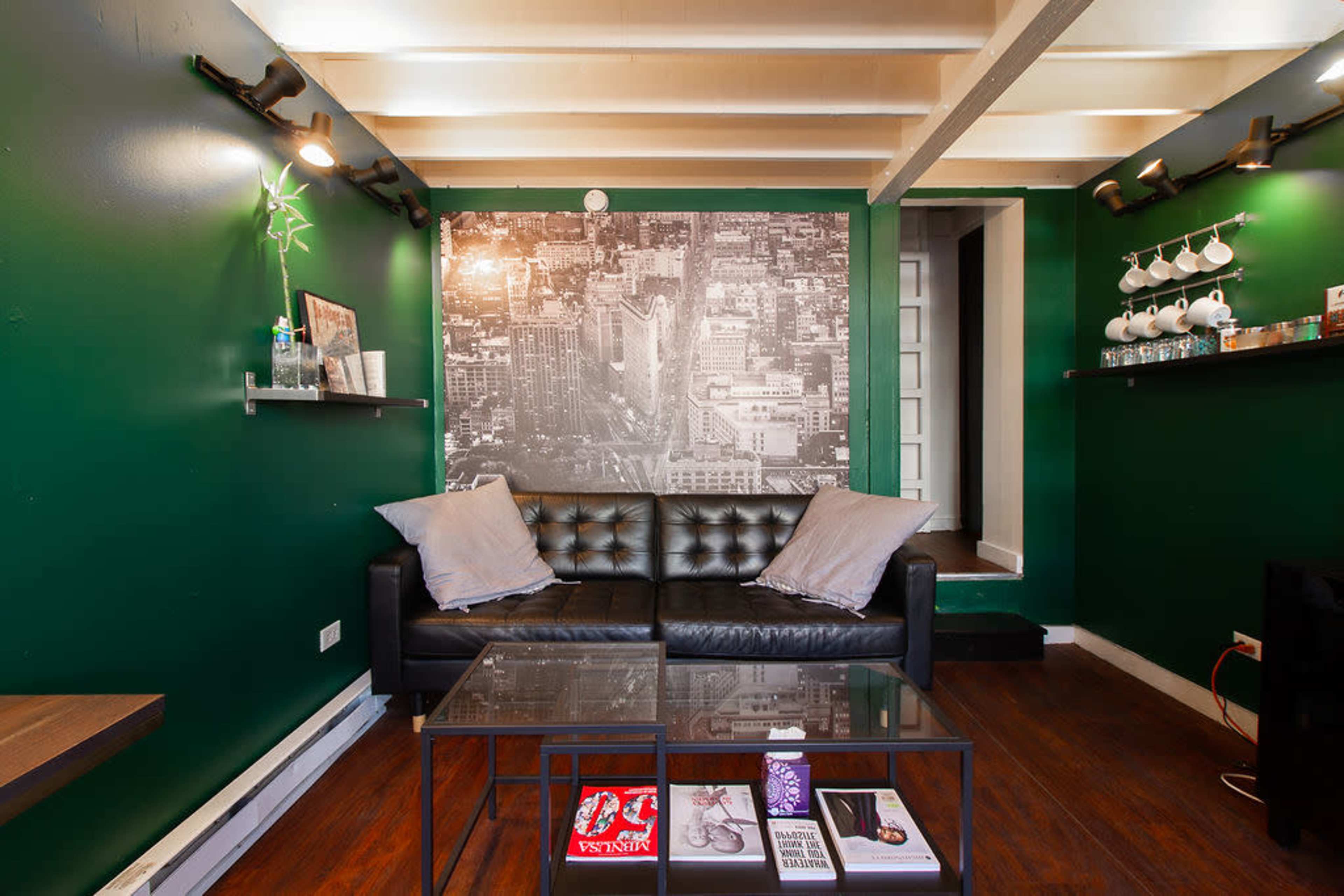 The image shows a small living space with dark green walls, a black leather couch, a coffee table with magazines, and shelves displaying various items.
