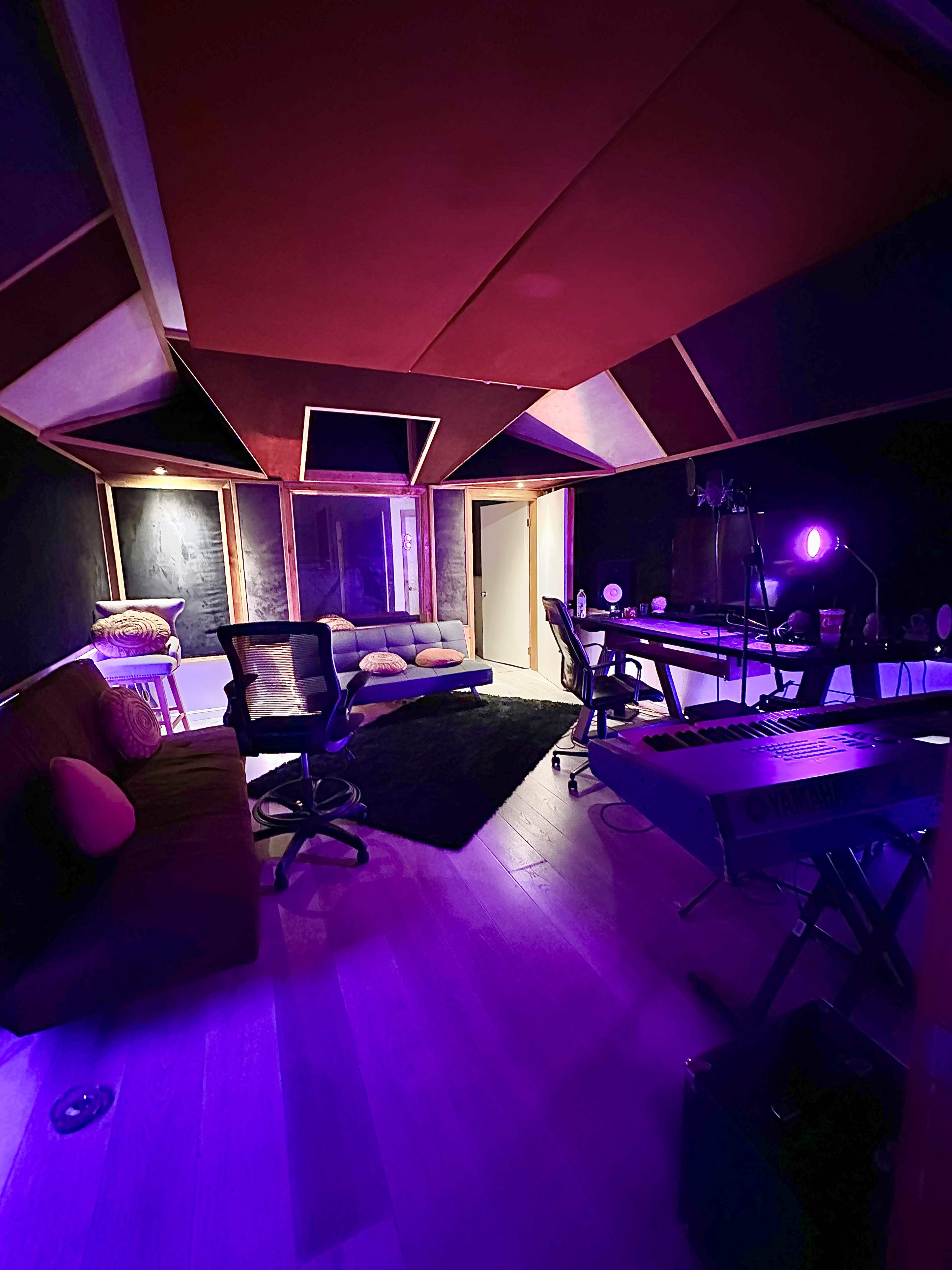 A music studio features a red and black color scheme, with a keyboard, chairs, and a sofa arranged around a central area illuminated by purple lighting.