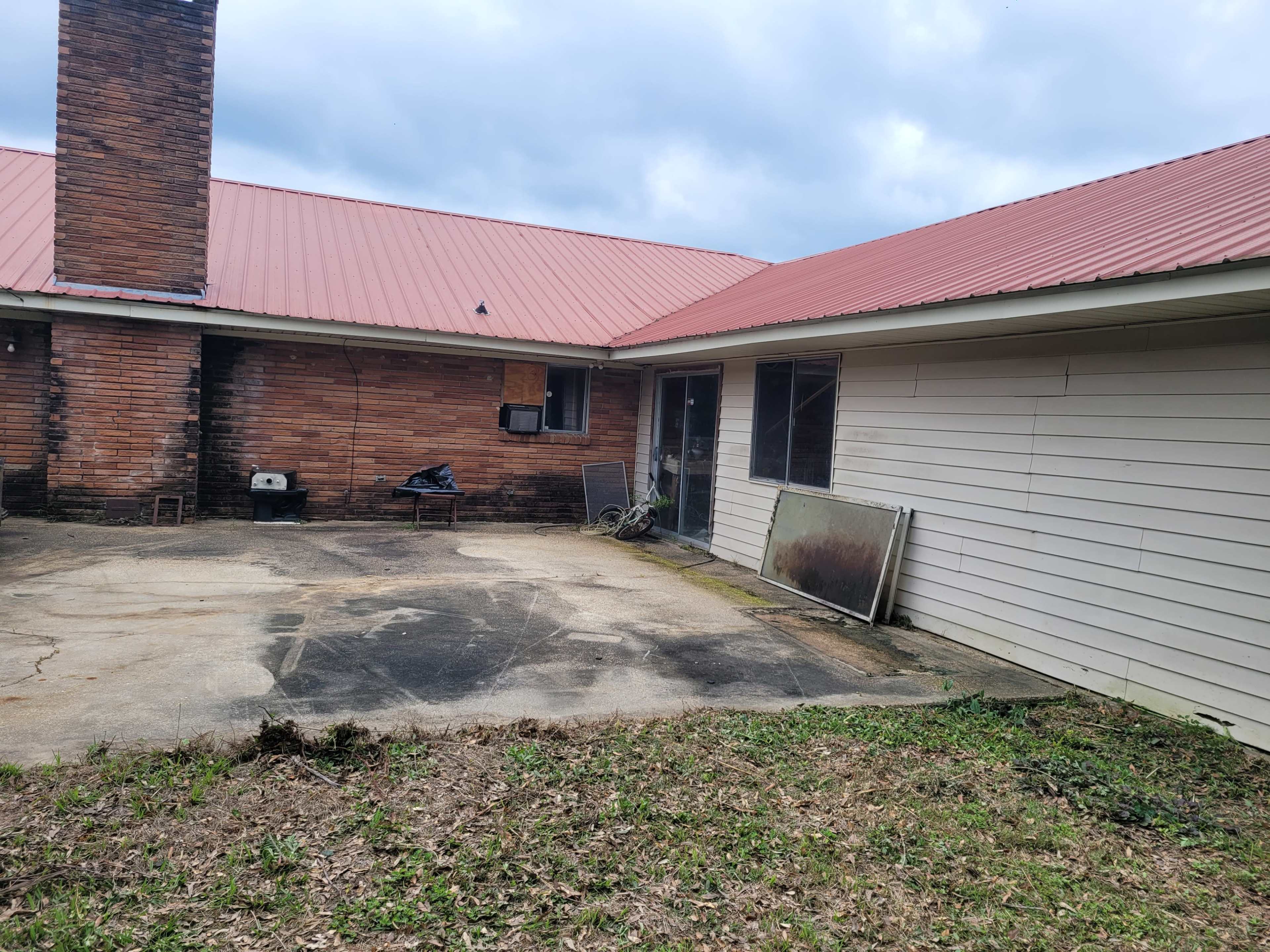 The image shows a backyard area of a house featuring a brick chimney, a red metal roof, a concrete patio, and some outdoor furniture and debris.