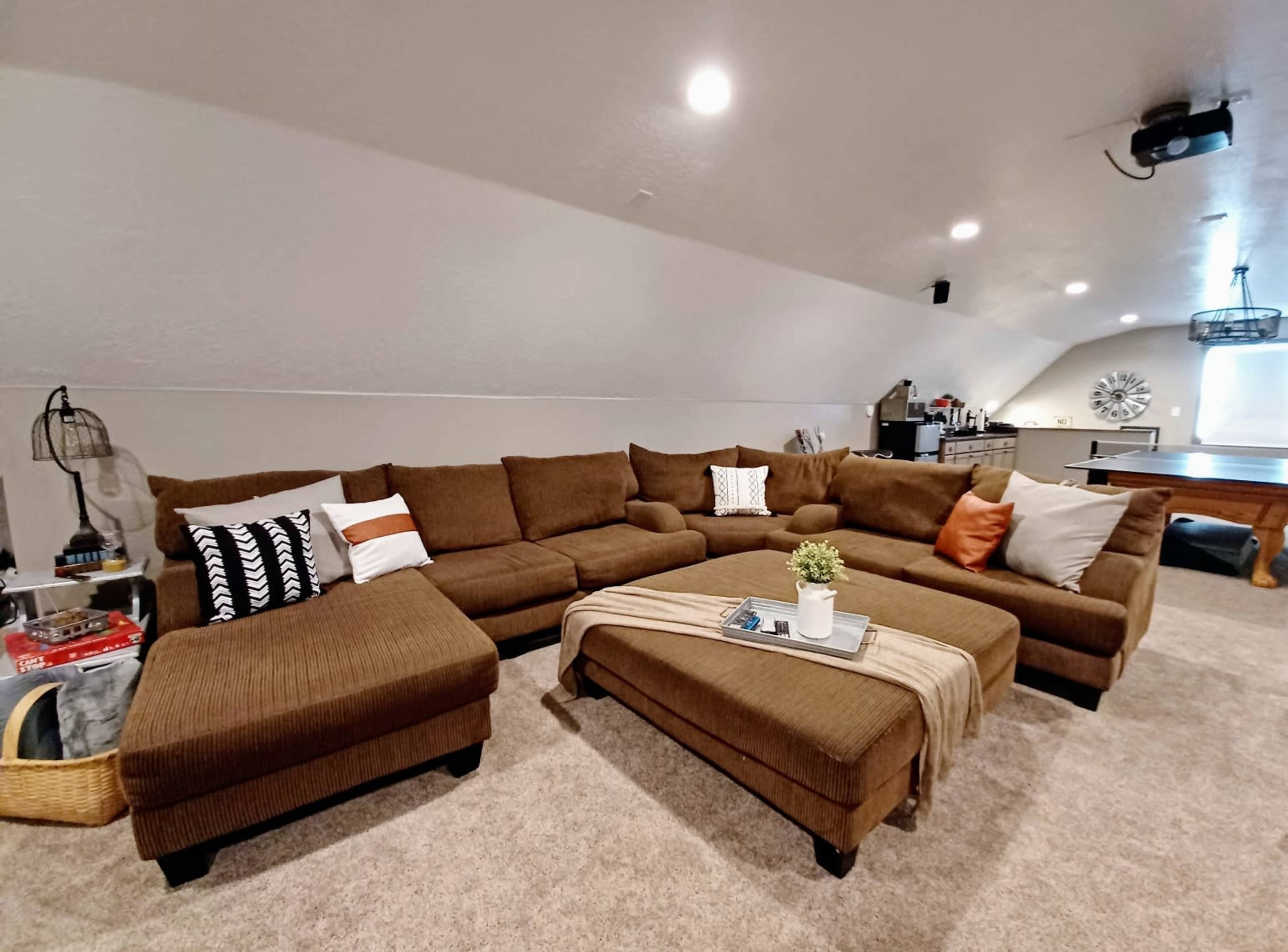 A spacious attic living room features a large brown sectional couch with a coffee table adorned with a plant and blanket, surrounded by neutral-colored walls and a pool table in the background.