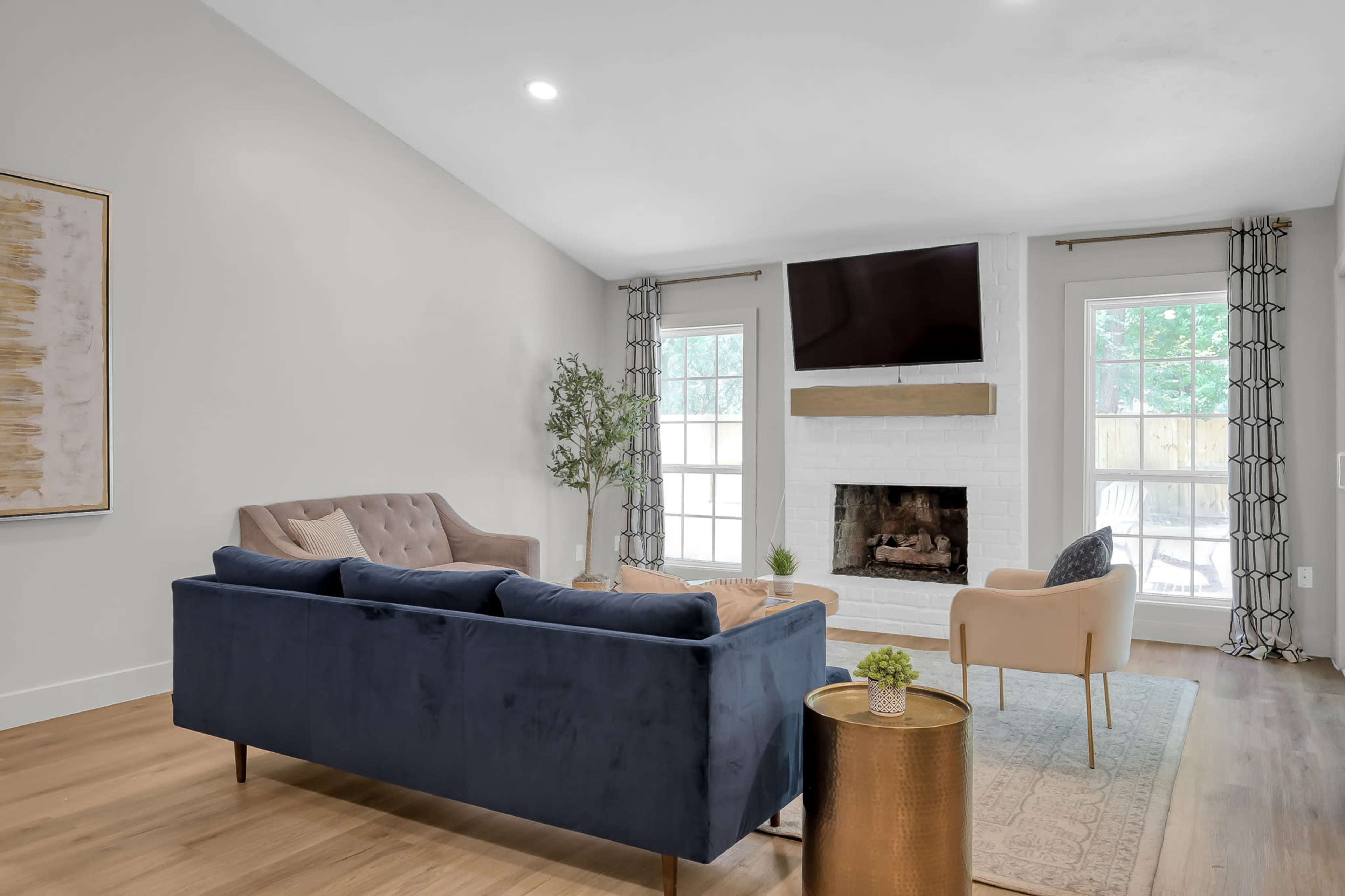 A modern living room features a blue velvet sofa, a light armchair, a wooden coffee table, and a white brick fireplace with a wooden mantel, all illuminated by natural light from large windows.