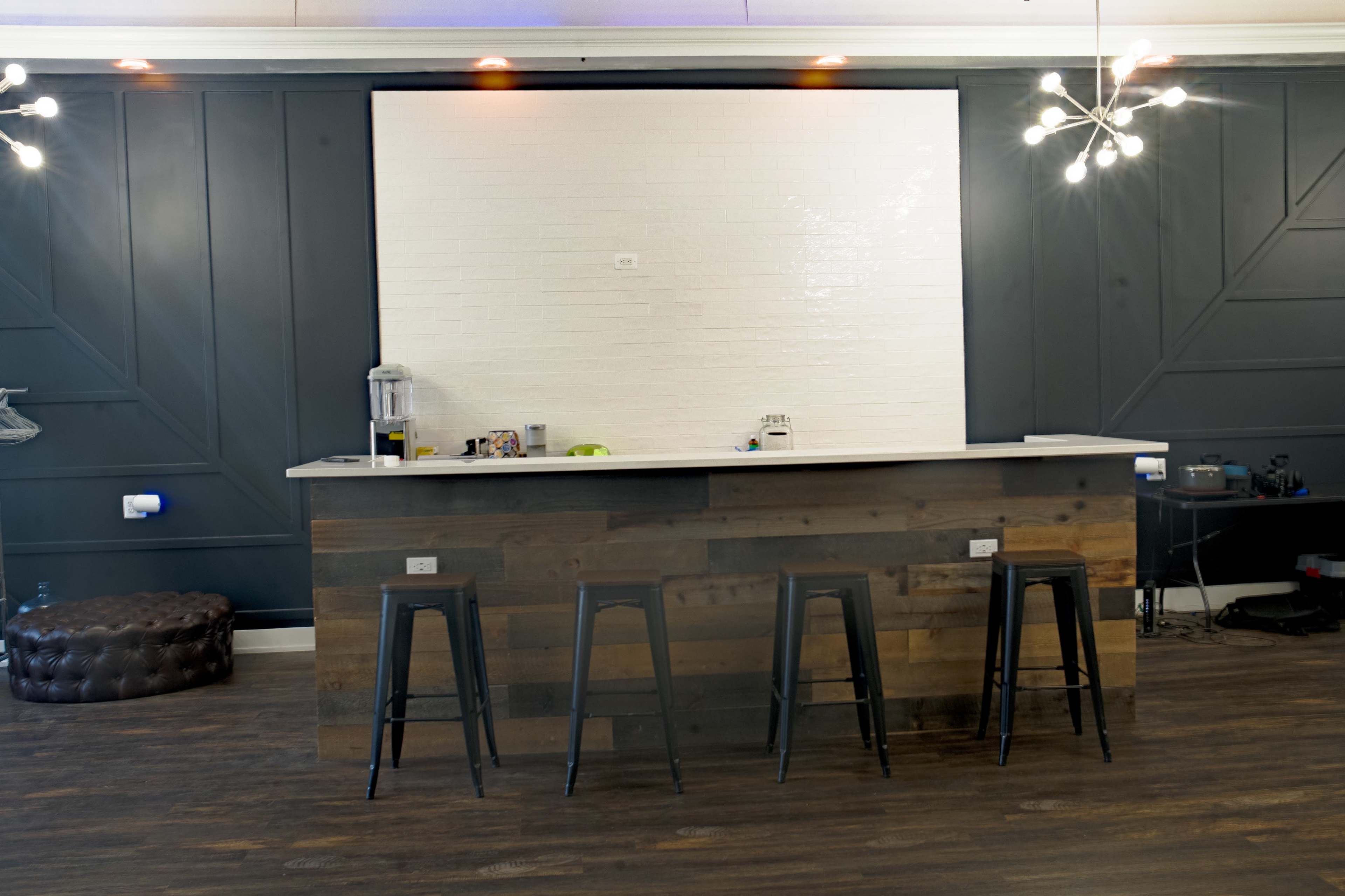 A minimalist bar area features a wooden counter with three black stools in front, set against a textured white wall and dark paneling.
