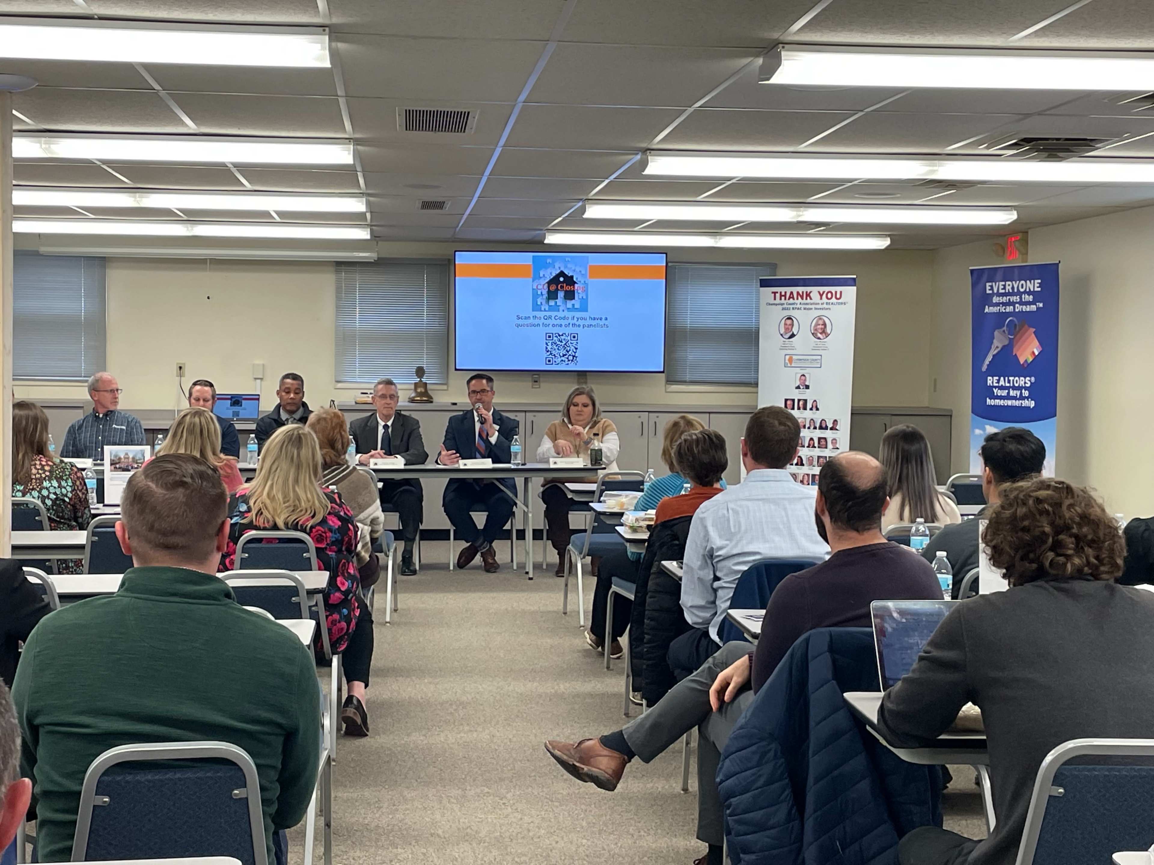 A panel of speakers discusses real estate topics in front of an audience seated at tables in a conference room.