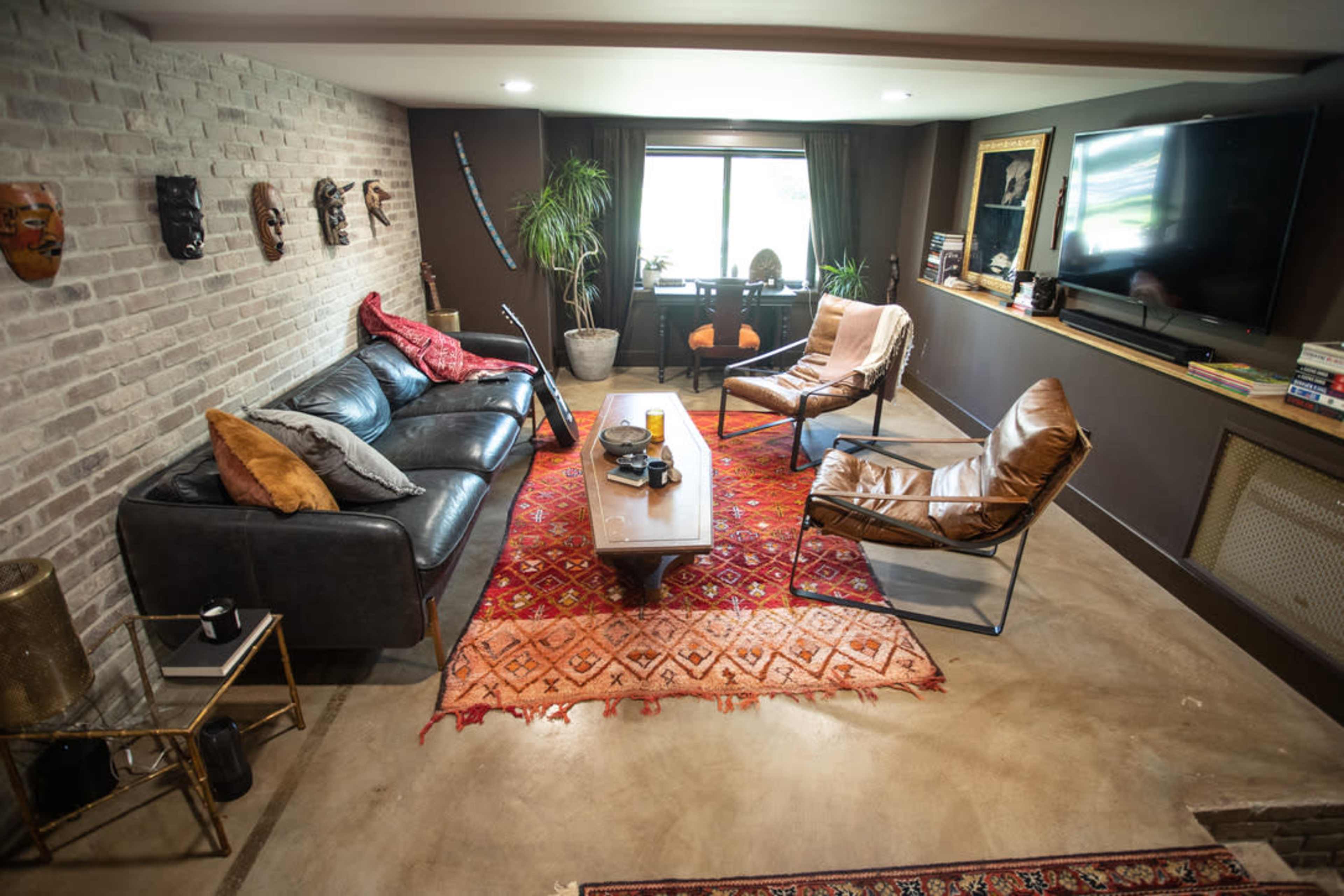 The image shows a modern living room featuring a black sofa, two leather chairs, a wooden coffee table on a patterned rug, and decorative masks on the brick wall.