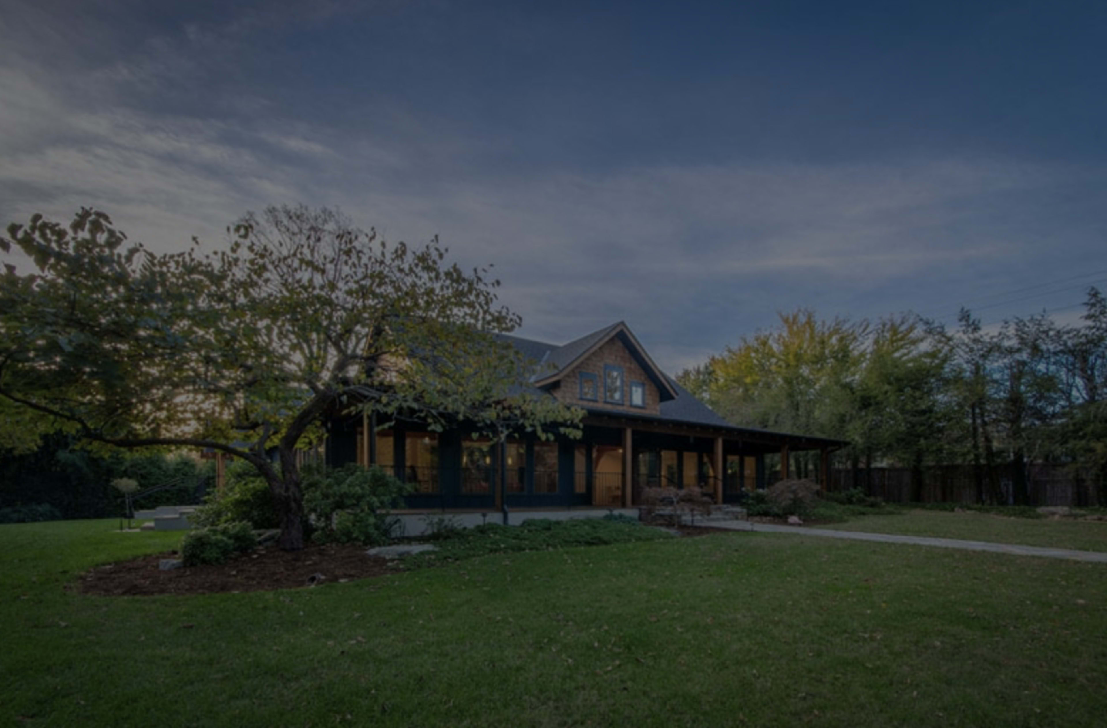A large house with a covered porch sits on a grassy lawn surrounded by trees under a cloudy sky.