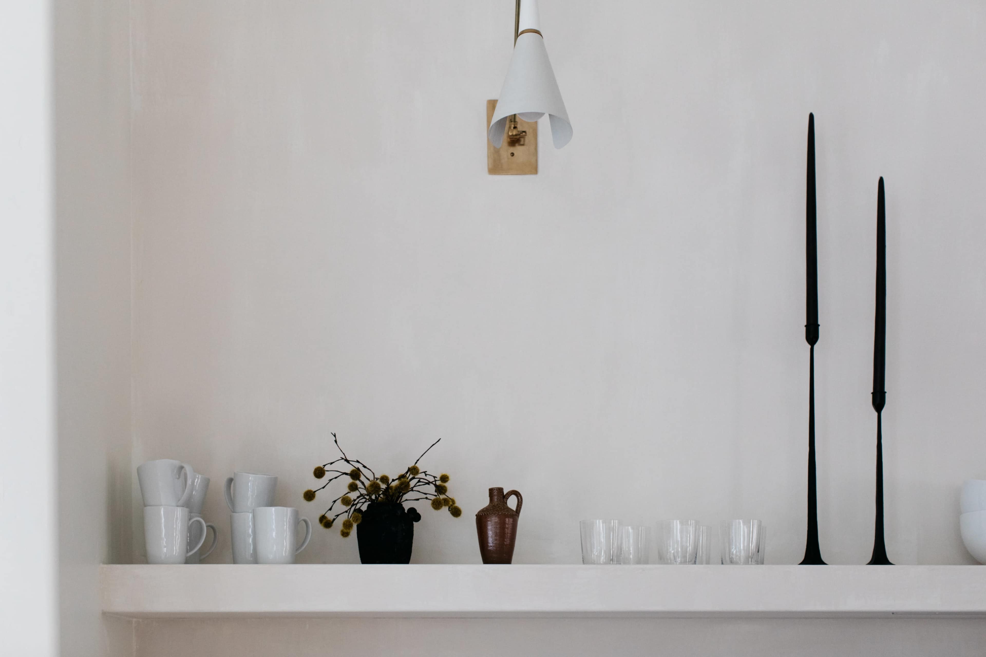 A shelf displays various ceramic mugs, a small vase with branches, a pitcher, and slender candle holders against a pale wall.