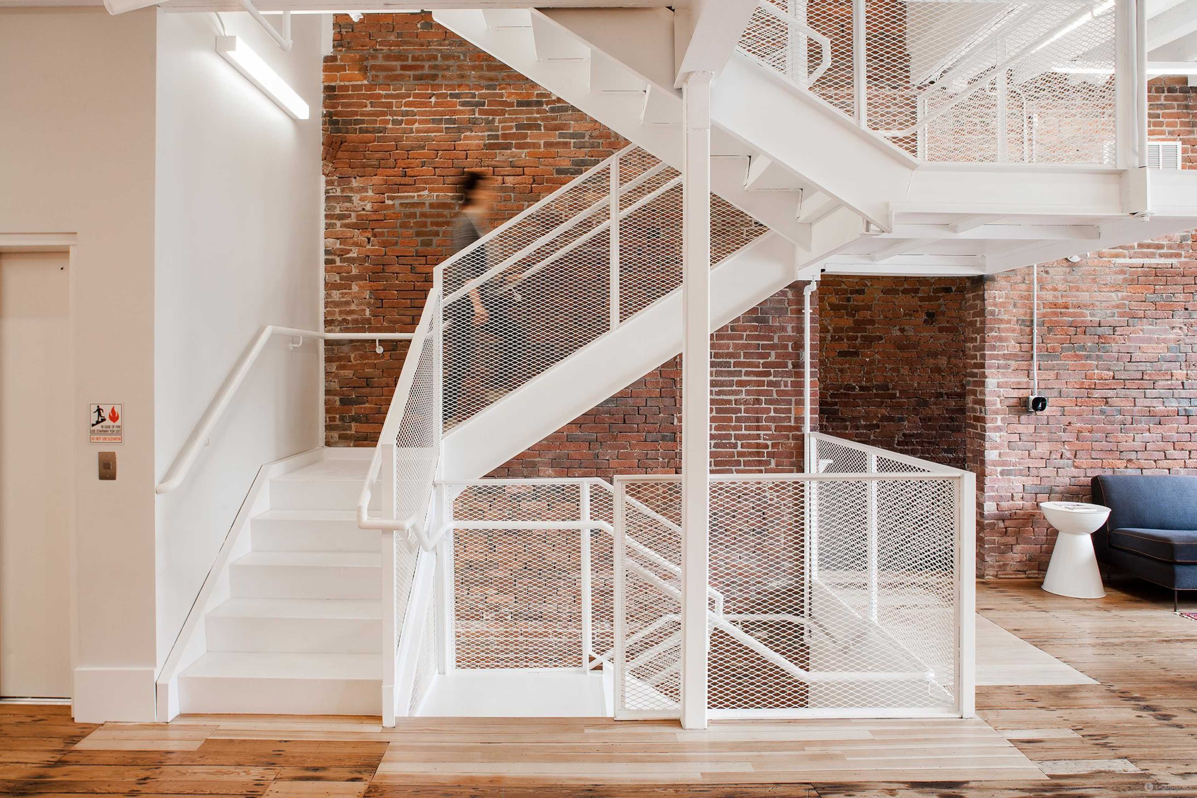 A person walks up a white, open staircase in a modern interior with exposed brick walls and wooden flooring.