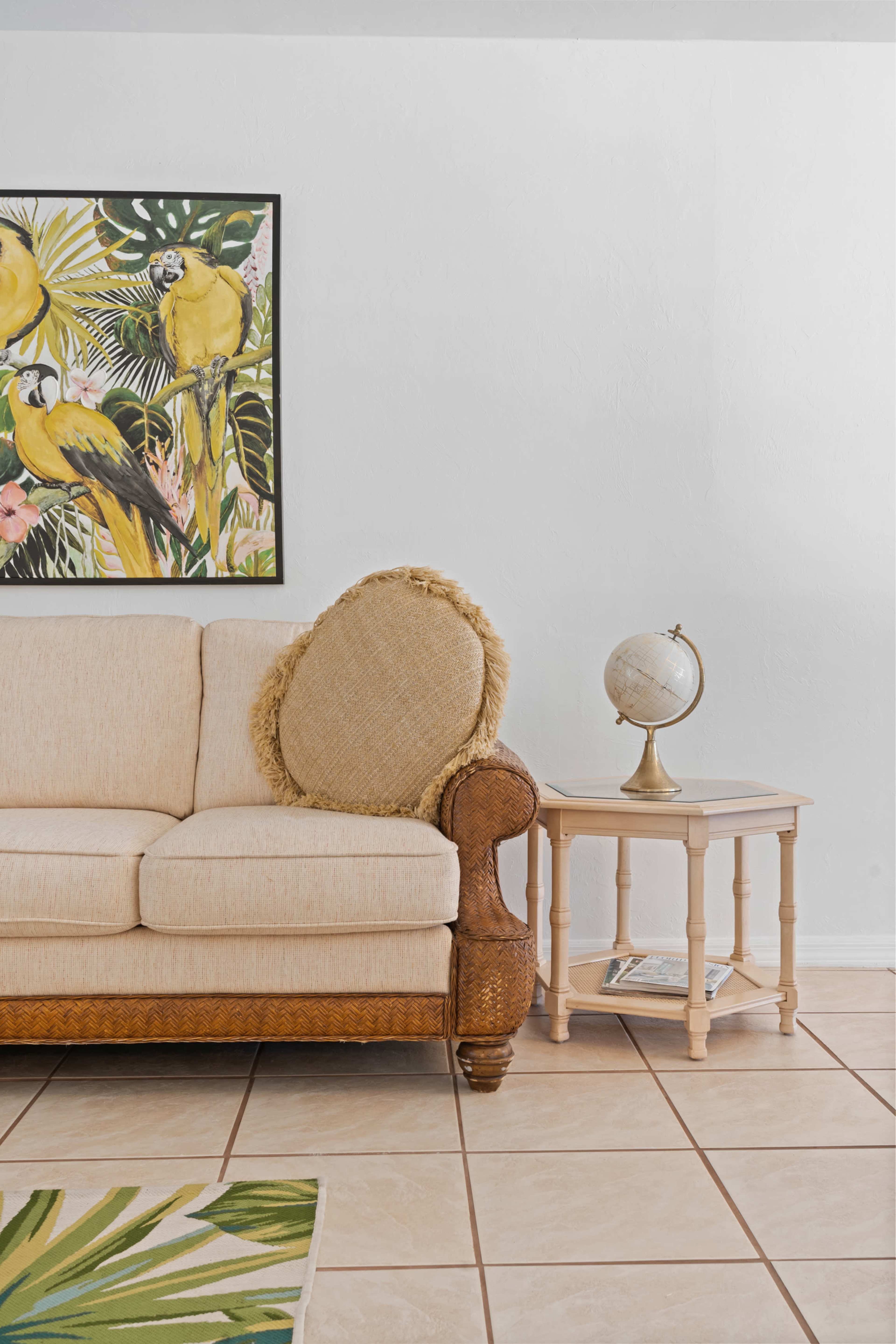 A beige sofa with a decorative pillow is positioned next to a wooden side table featuring a globe, against a backdrop of a tropical-themed wall art.