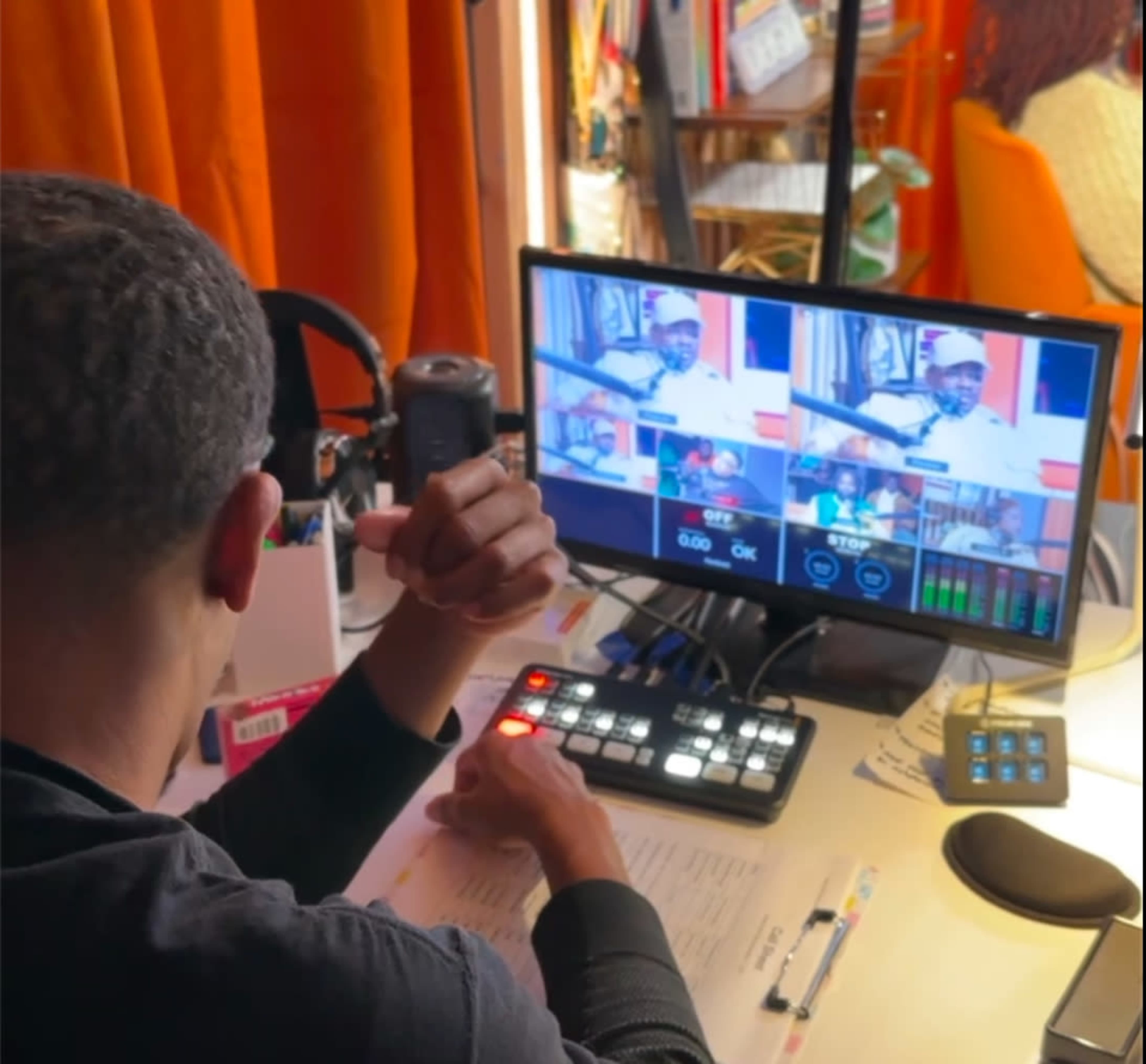 A person is sitting at a desk, monitoring a video feed on a screen while using a control panel to manage audio and visuals.