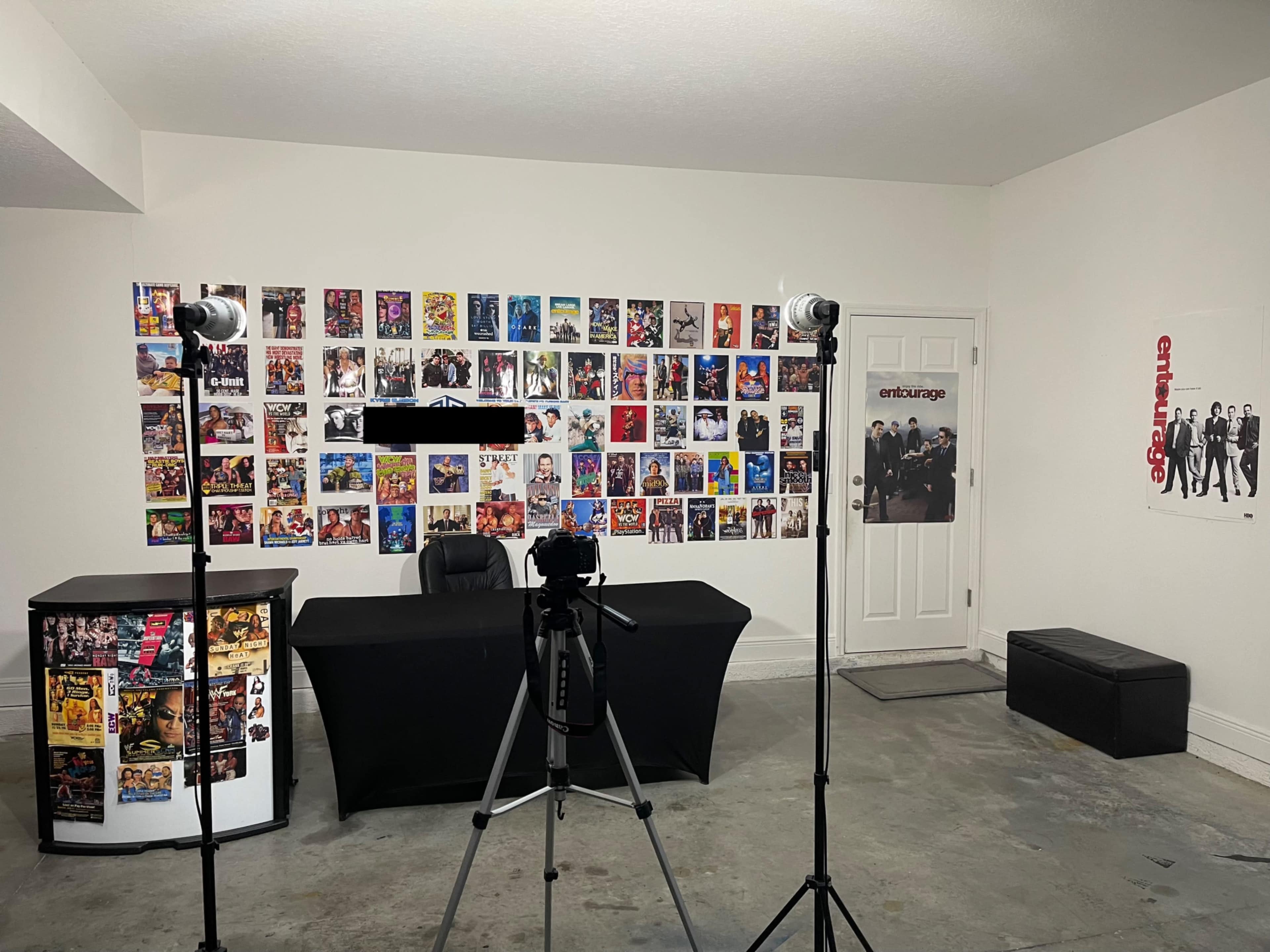 The image shows a minimalist room featuring a table with a black chair and several photography lights, surrounded by walls covered with colorful movie posters and a door leading to another area.