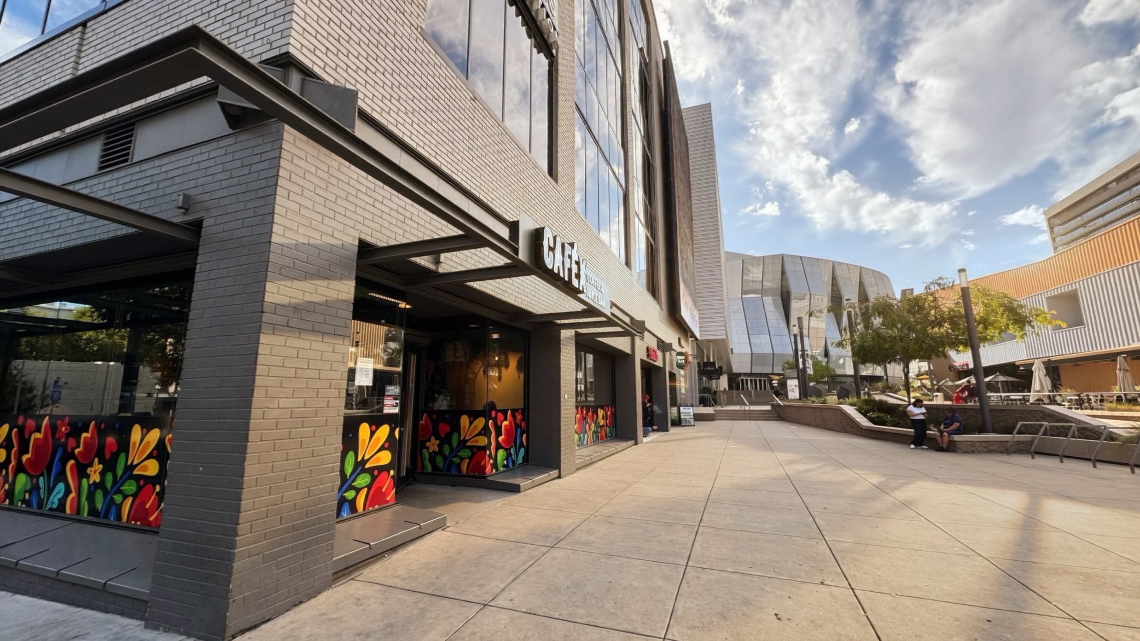 The image shows a modern building with a colorful mural on its entrance, situated near an outdoor seating area and another contemporary structure in the background.