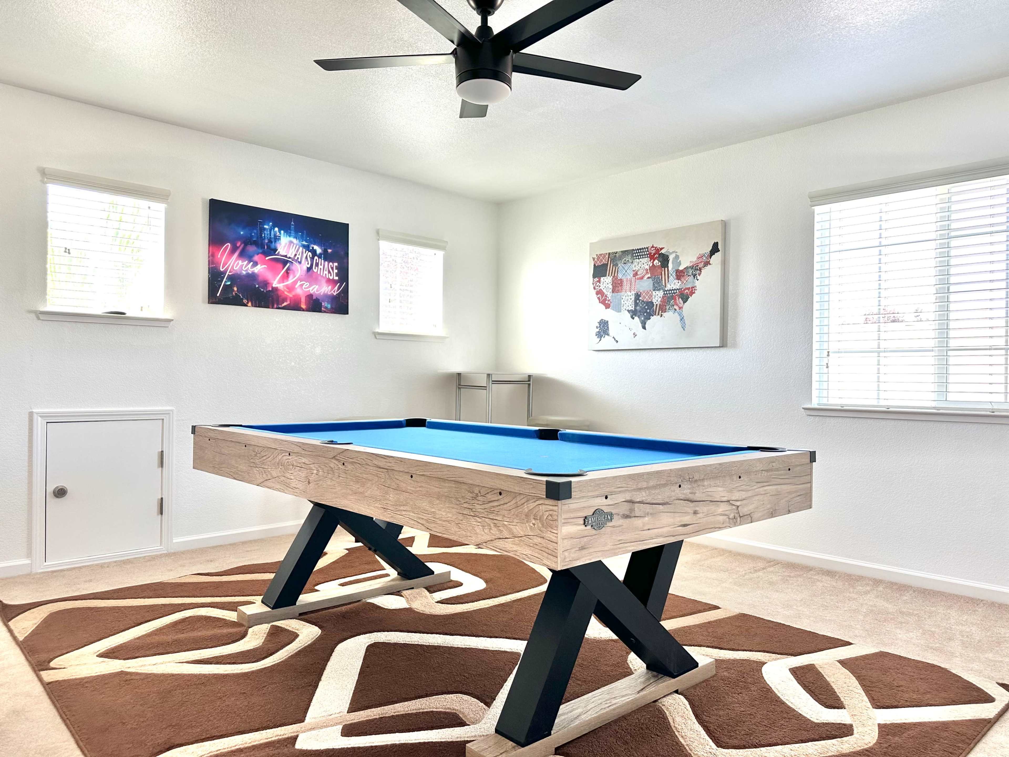 The image shows a game room featuring a blue pool table with a wooden frame, situated between two windows and decorated with framed art on the walls.