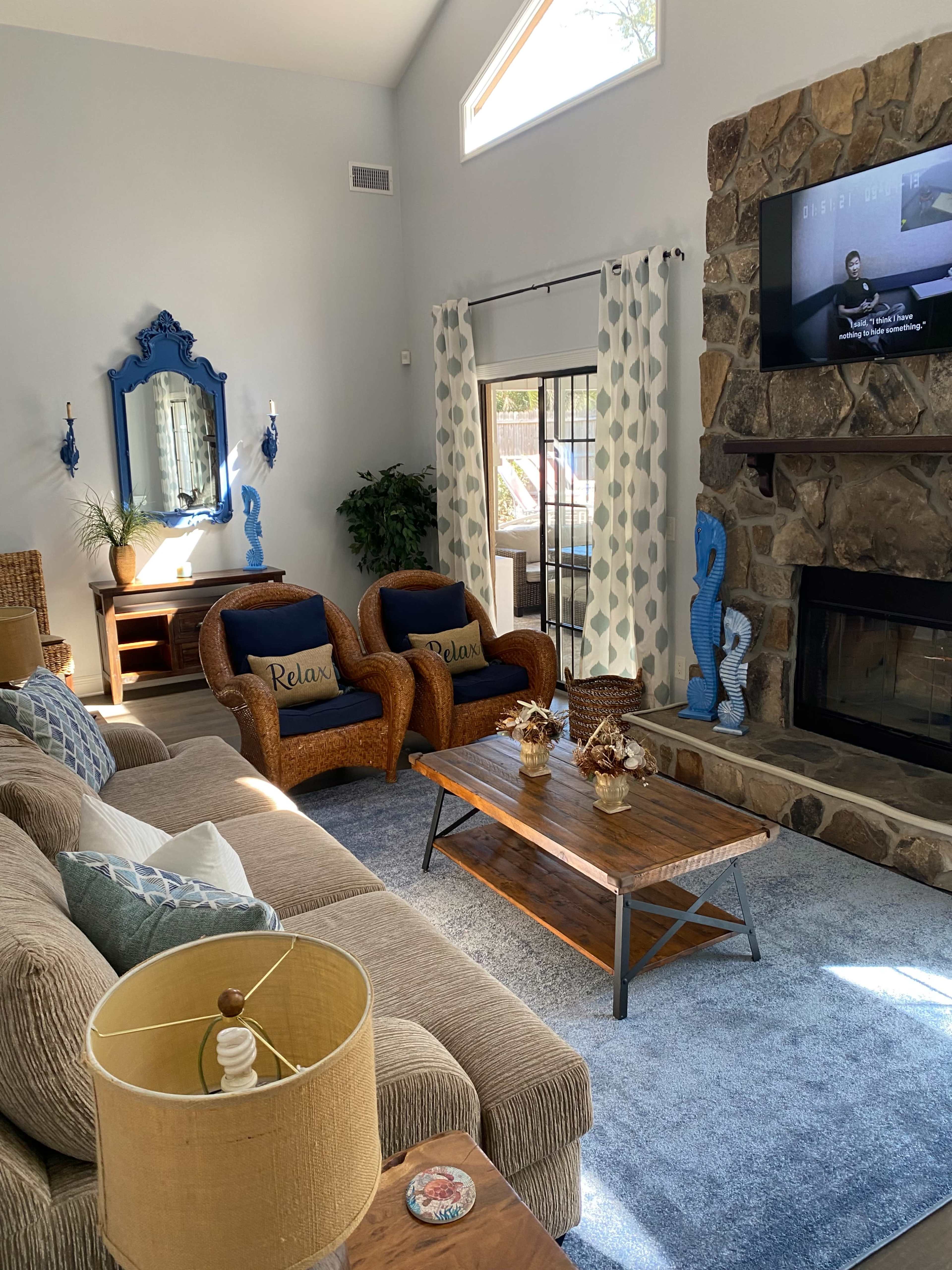 A cozy living room features a brown sectional sofa, two blue chairs with "Relax" cushions, a wooden coffee table, and a stone fireplace, with a large window allowing natural light to enter.