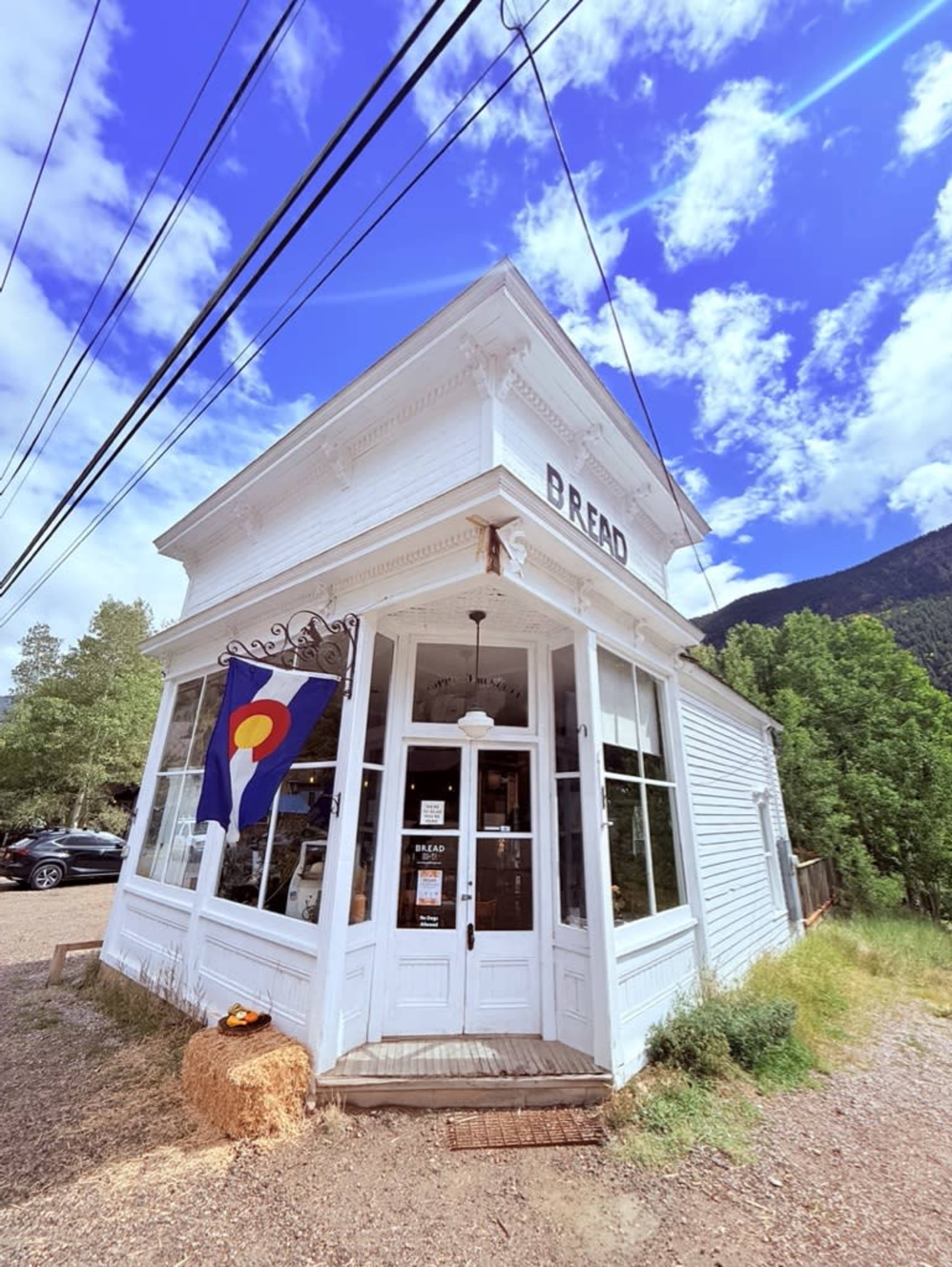 The image shows a quaint white bakery building with large windows and a Colorado state flag hanging outside, set against a backdrop of blue skies and greenery.