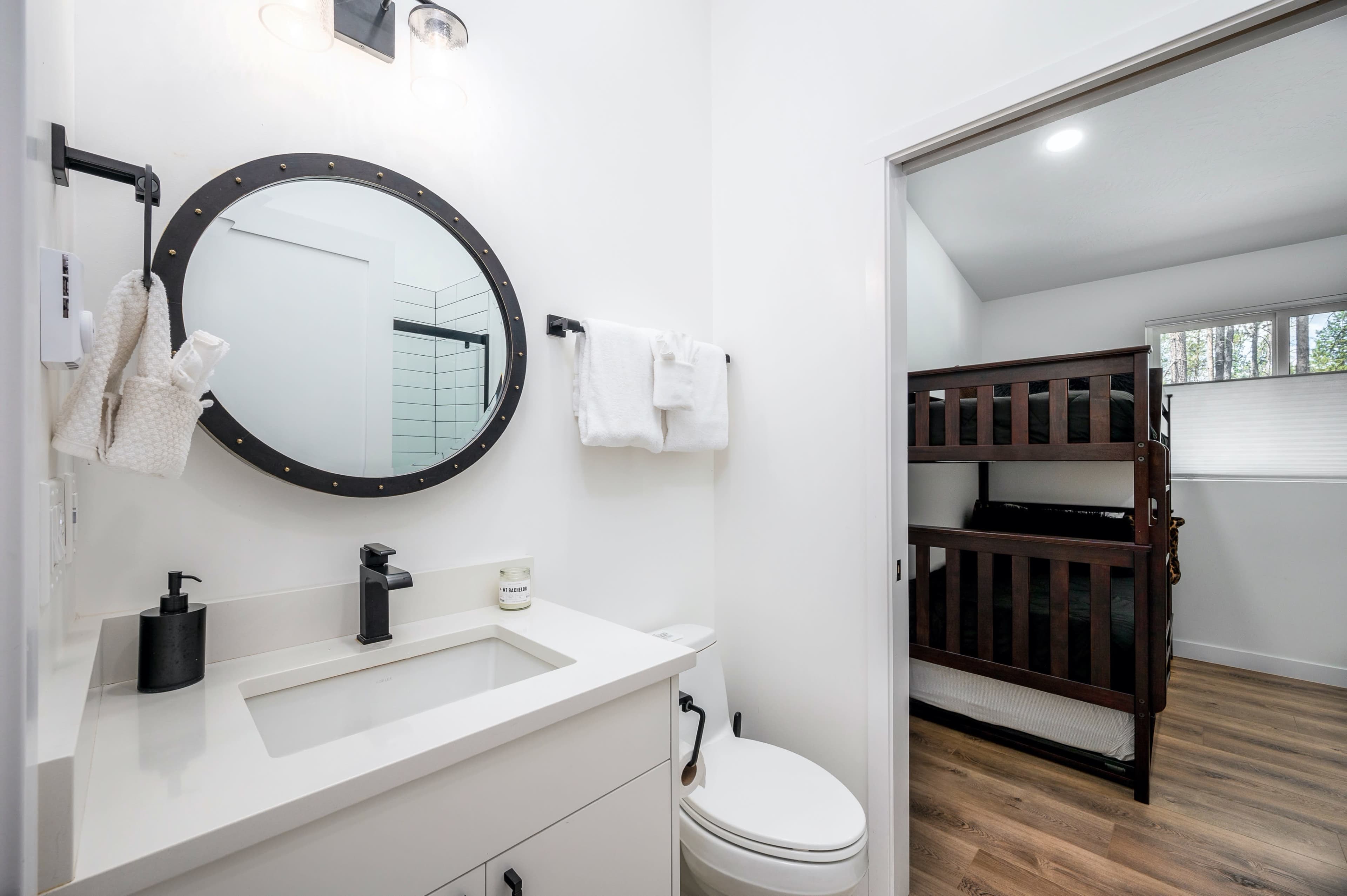 A modern bathroom featuring a circular mirror above a white sink, a towel rack, and a toilet, with a bunk bed visible in the adjacent room.