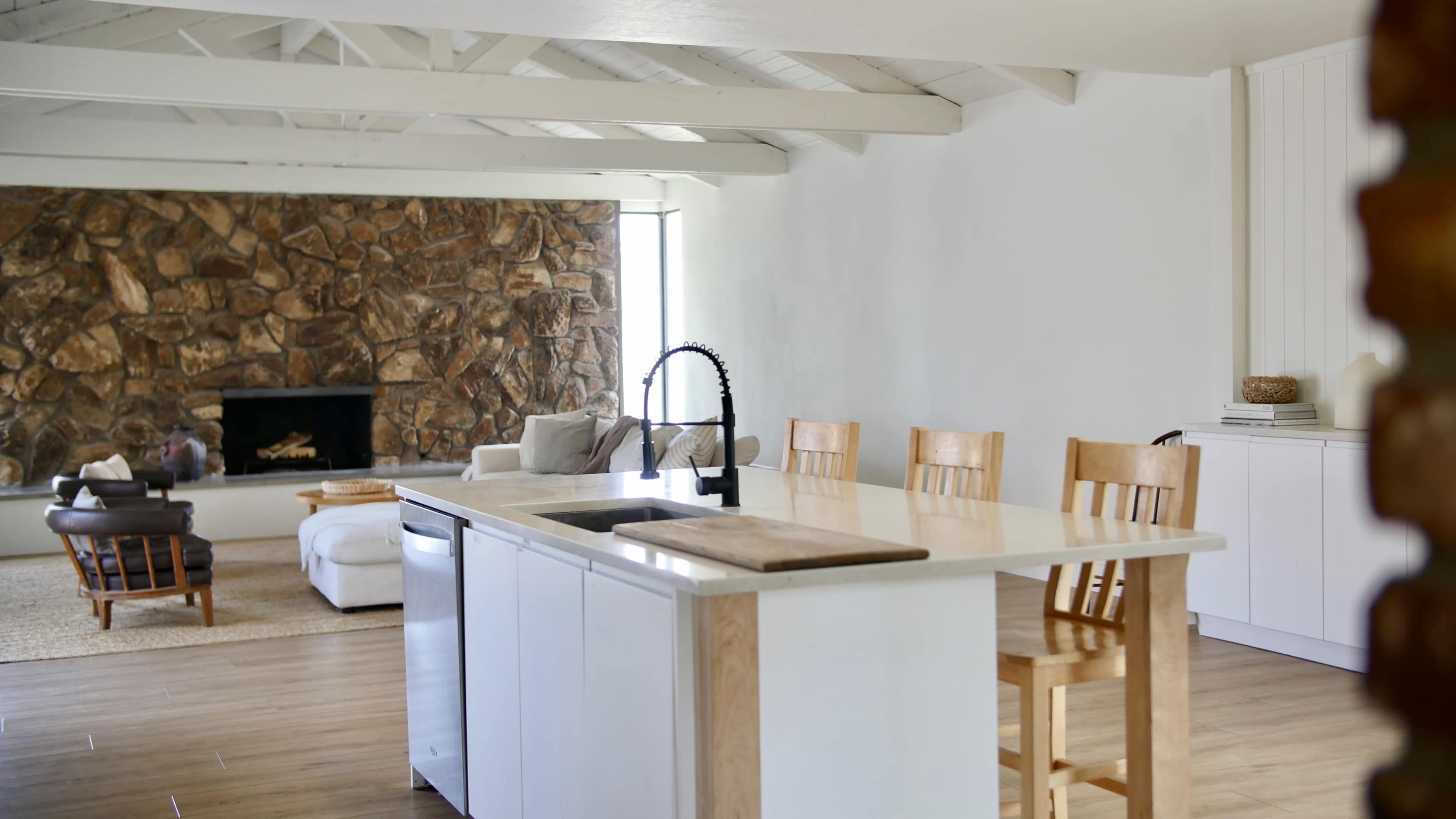 A modern kitchen features a central island with a sink and three wooden barstools, adjacent to a living area with a stone accent wall and a plush sofa.