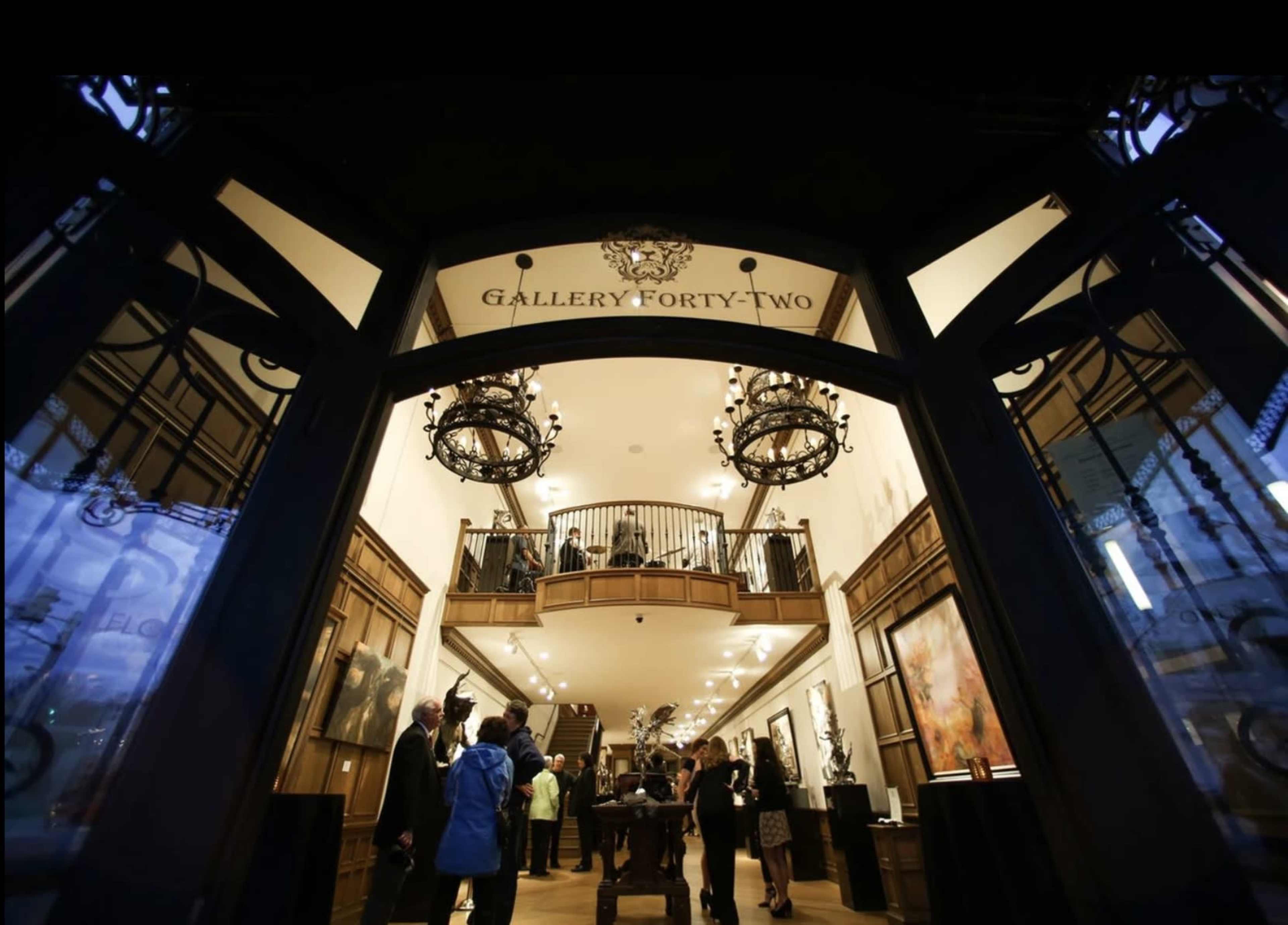 A well-lit art gallery entrance features a staircase leading to an upper level, with people mingling inside.