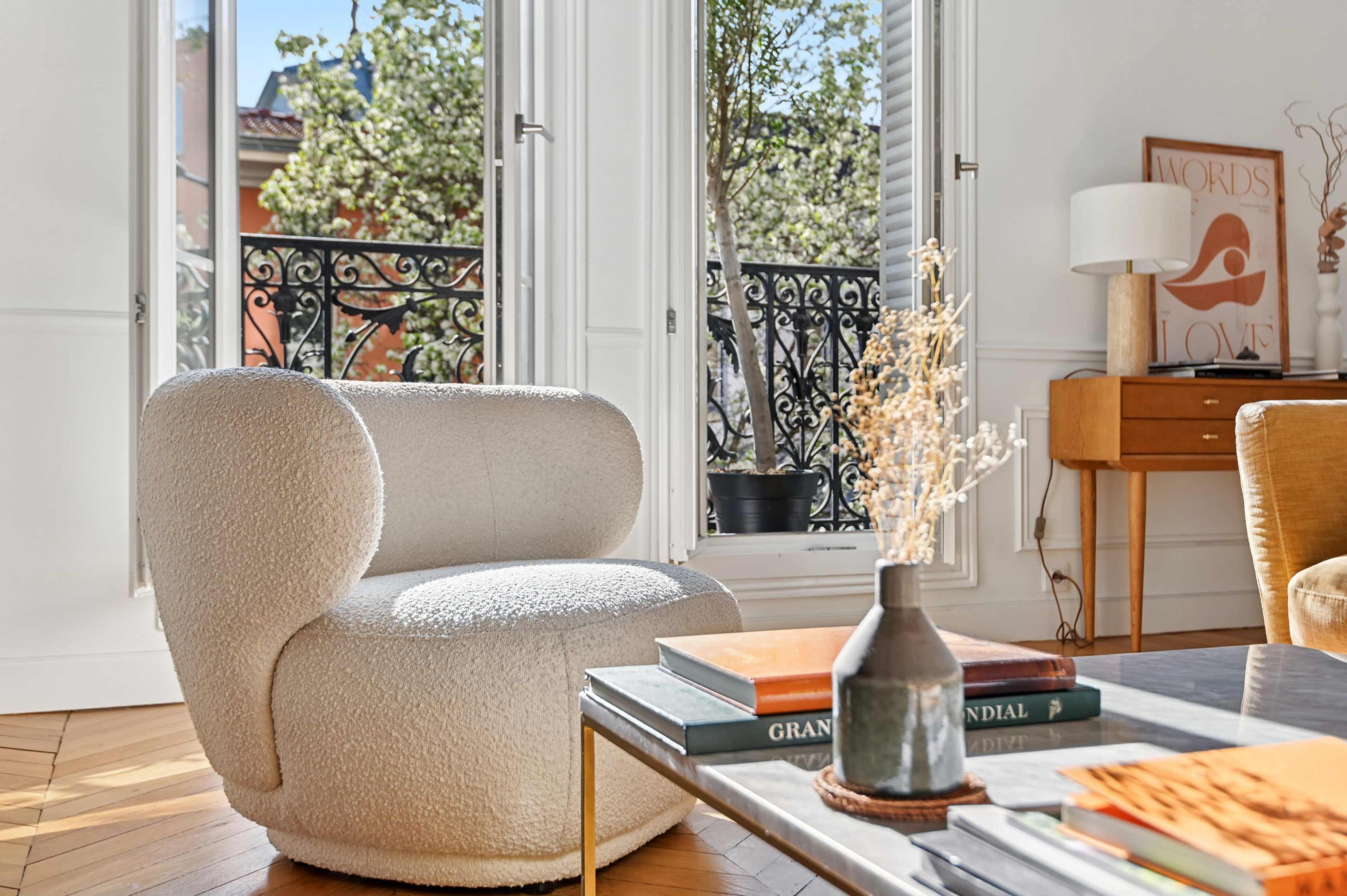 A round, white chair is placed next to a coffee table with books and a vase, while large windows offer a view of a balcony with decorative ironwork.