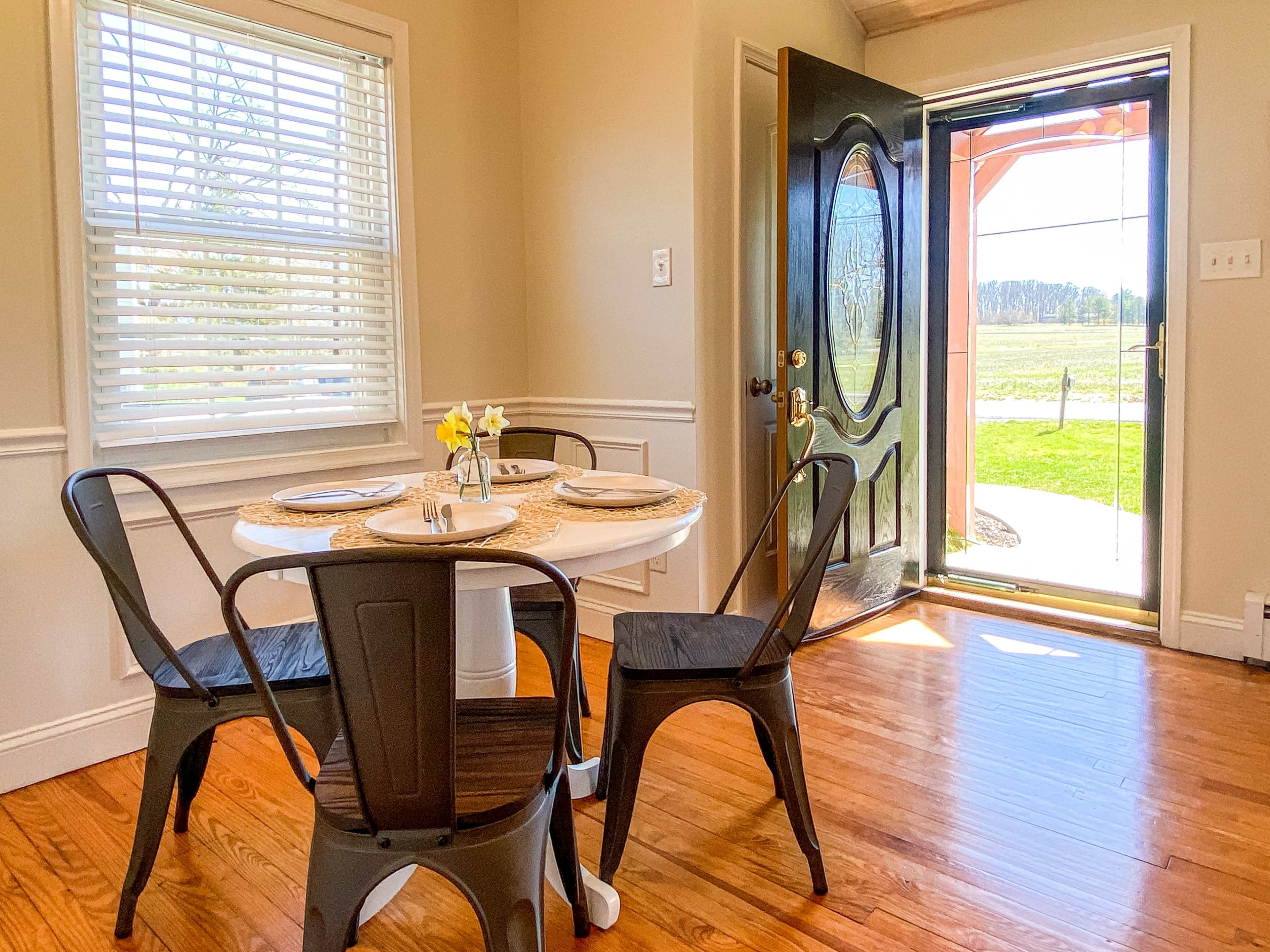 A small dining area features a round table with place settings and four black chairs, adjacent to an open door leading to an outdoor view.