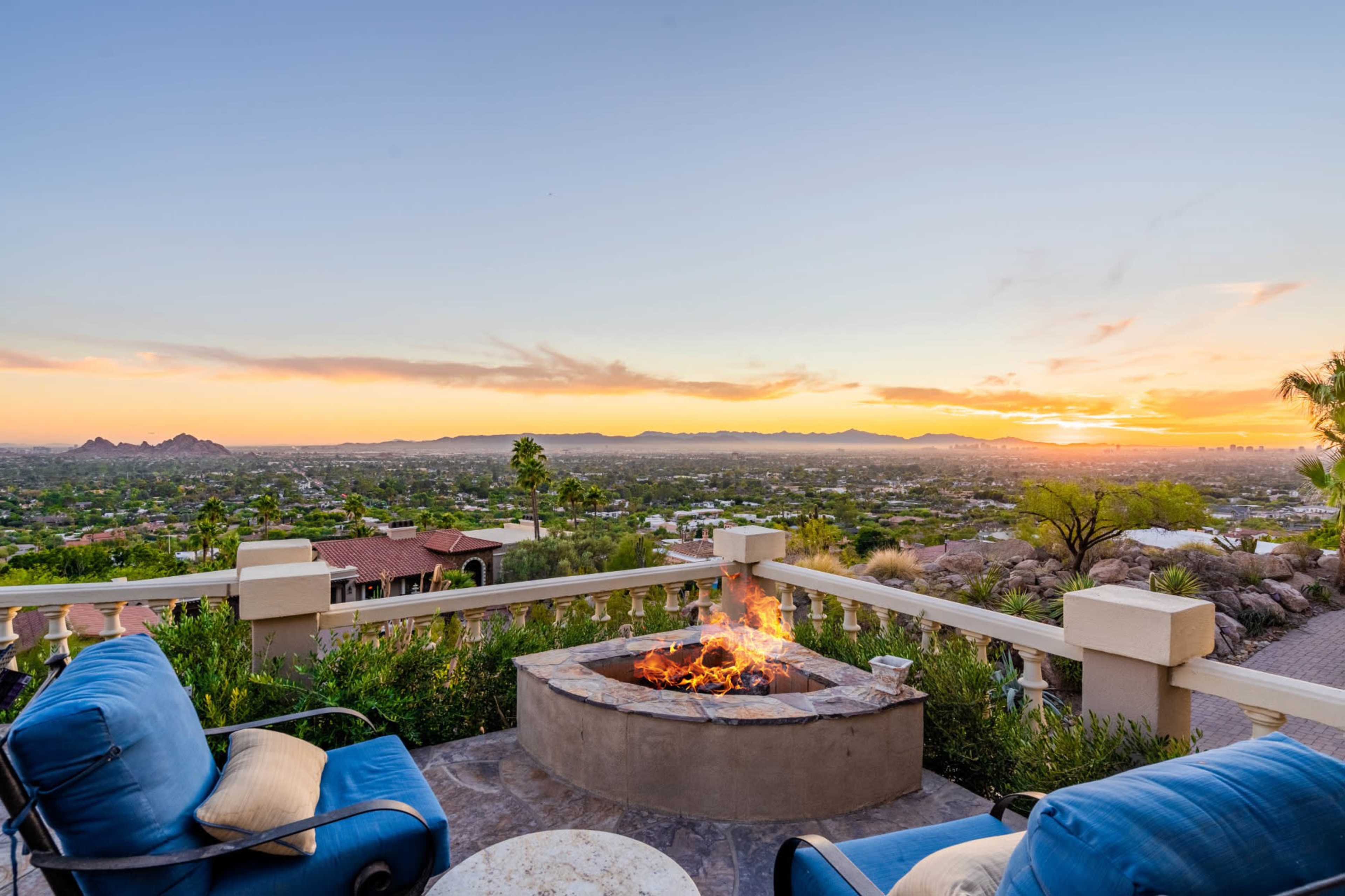 A fire pit surrounded by blue chairs offers a view of a sunset over a distant landscape and homes.